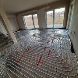 Rozłożona instalacja ogrzewania podłogowego na czerwonej siatce w surowym wnętrzu domu w trakcie budowy, widoczne okna i schody.