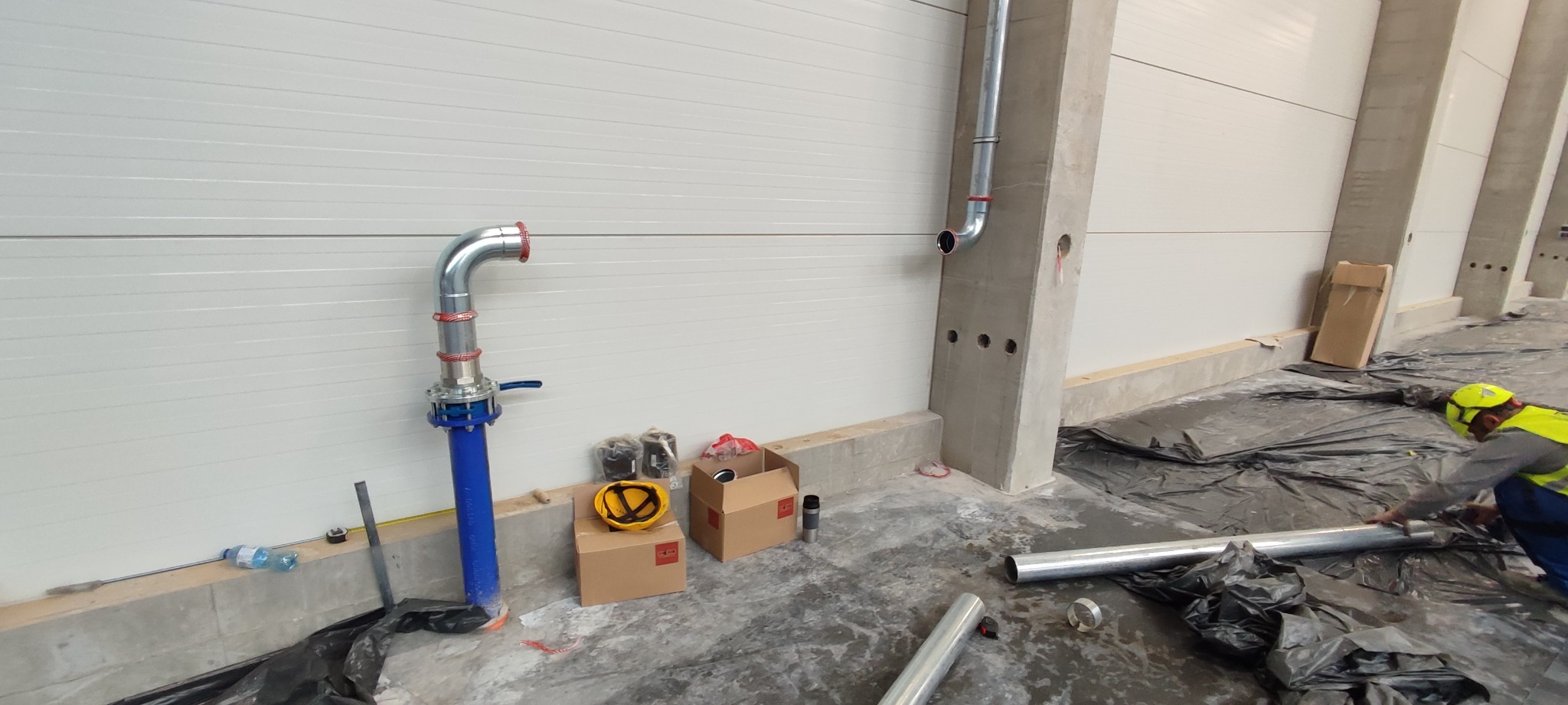 Montaż rur w hali produkcyjnej: pionowa, niebieska rura z zaworem, srebrne kolanko, materiały montażowe w kartonach, pracownik w żółtym kasku przy srebrnych rurach na folii ochronnej, betonowe filary.