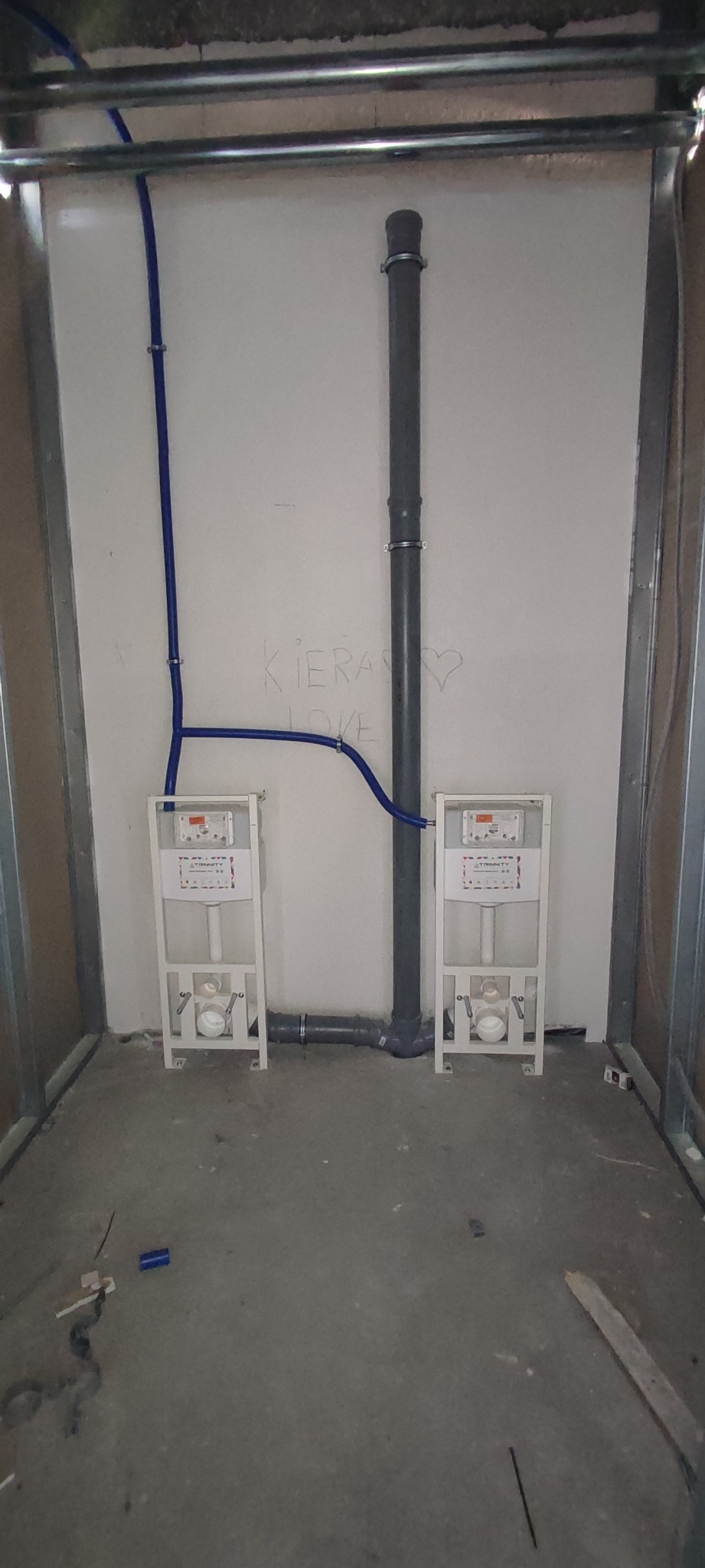 Dwa stelaże podtynkowe WC w trakcie montażu, z podłączonymi niebieskimi rurami doprowadzającymi wodę i szarą rurą odpływową, widoczne w surowym wnętrzu z metalową konstrukcją ścian działowych...