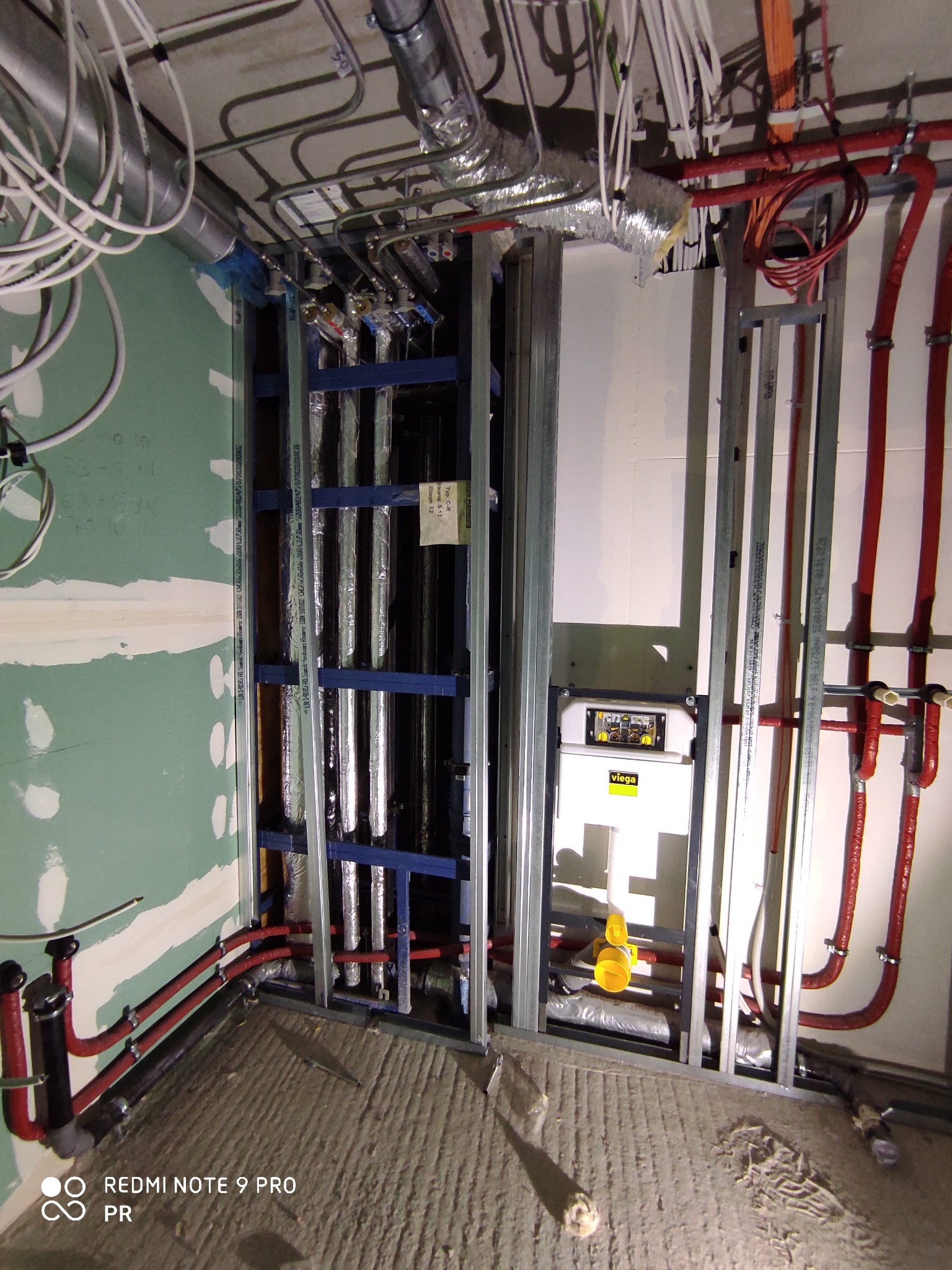 Fragment instalacji w surowym stanie: rury grzewcze w izolacji, metalowa konstrukcja szkieletowa, biały zbiornik Viega, liczne kable i przewody pod sufitem, ściany w trakcie wykańczania...