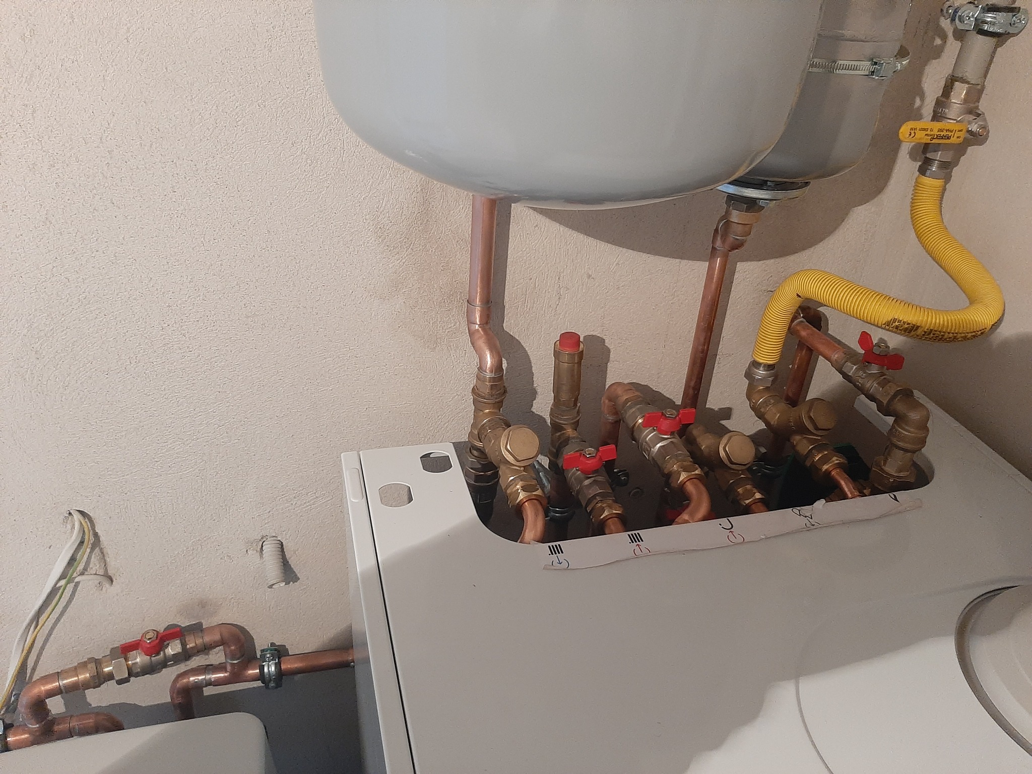 Szczegółowe ujęcie kotła grzewczego z widocznymi miedzianymi rurami, zaworami z czerwonymi pokrętłami i żółtym elastycznym przewodem gazowym, zamontowanego przy kremowej ścianie.