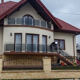 STAL-INOX - Dwupiętrowy dom z czerwonym dachem i balkonami z przezroczystymi, szklanymi balustradami, na których pracuje osoba w czarnym ubraniu, widoczna zza metalowego ogrodzenia z kamienną podmurówką.