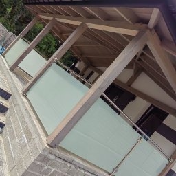 STAL-INOX - Zadaszony balkon z drewnianą konstrukcją i matowymi szklanymi panelami, widok z dołu na elewację budynku.