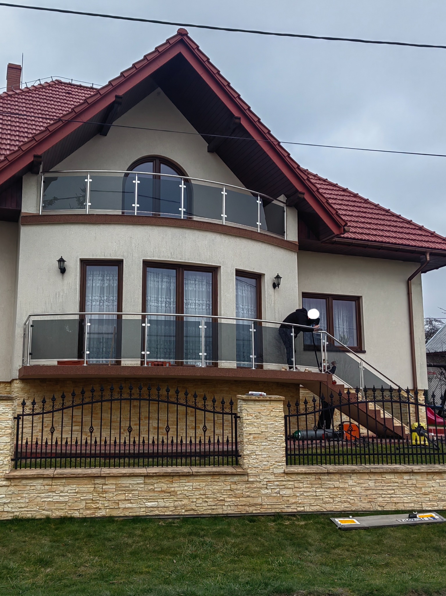 Dwupiętrowy dom z czerwonym dachem i balkonami z przezroczystymi, szklanymi balustradami, na których pracuje osoba w czarnym ubraniu, widoczna zza metalowego ogrodzenia z kamienną podmurówką.