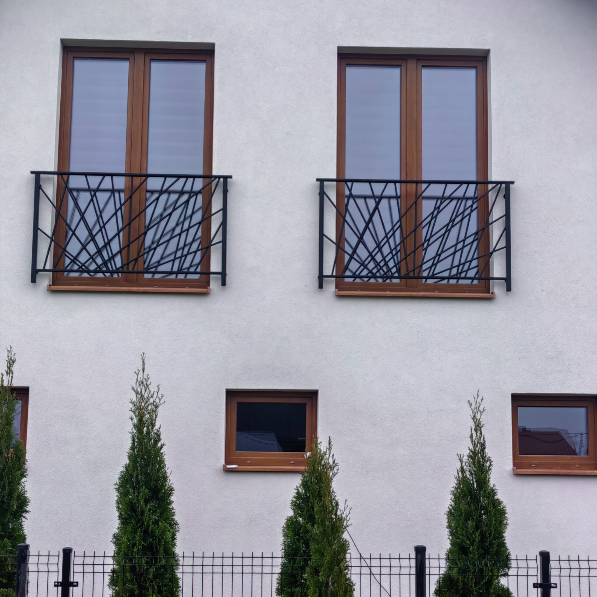 Elewacja budynku z dwoma oknami balkonowymi zabezpieczonymi czarną, geometryczną konstrukcją metalową. Na dole widoczne mniejsze okna i tuje za metalowym ogrodzeniem.