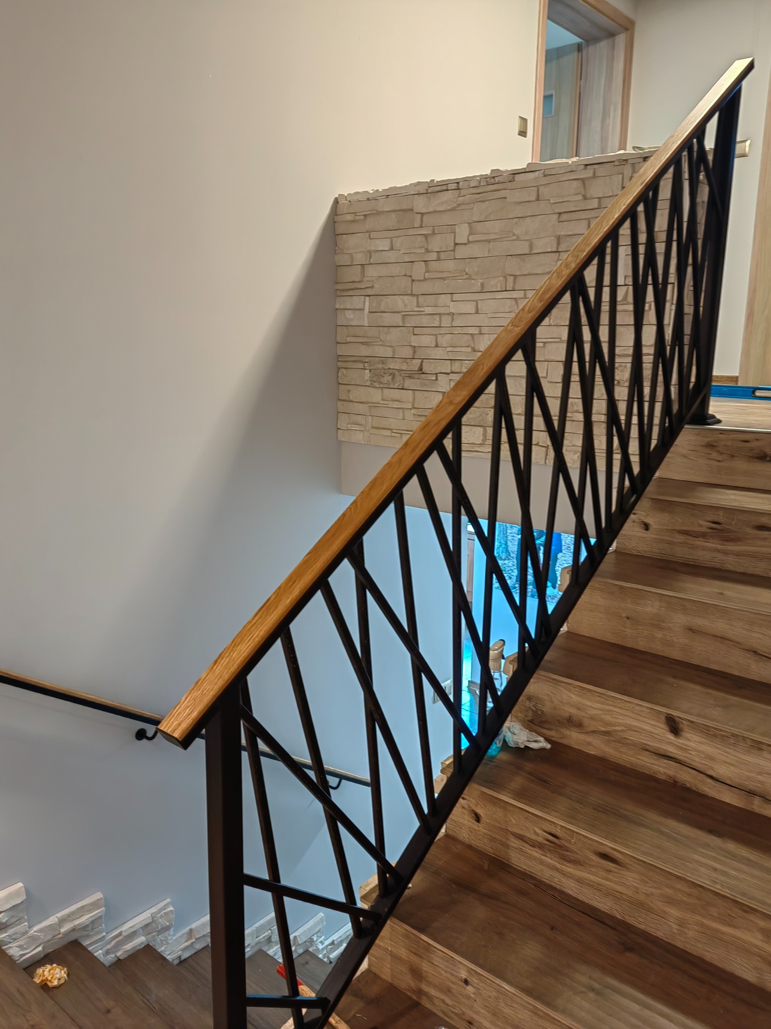 Schody drewniane z metalową balustradą o geometrycznym wzorze rombów, drewniany pochwyt, widoczne fragmenty ściany wykończonej kamieniem dekoracyjnym i drewniane stopnie.