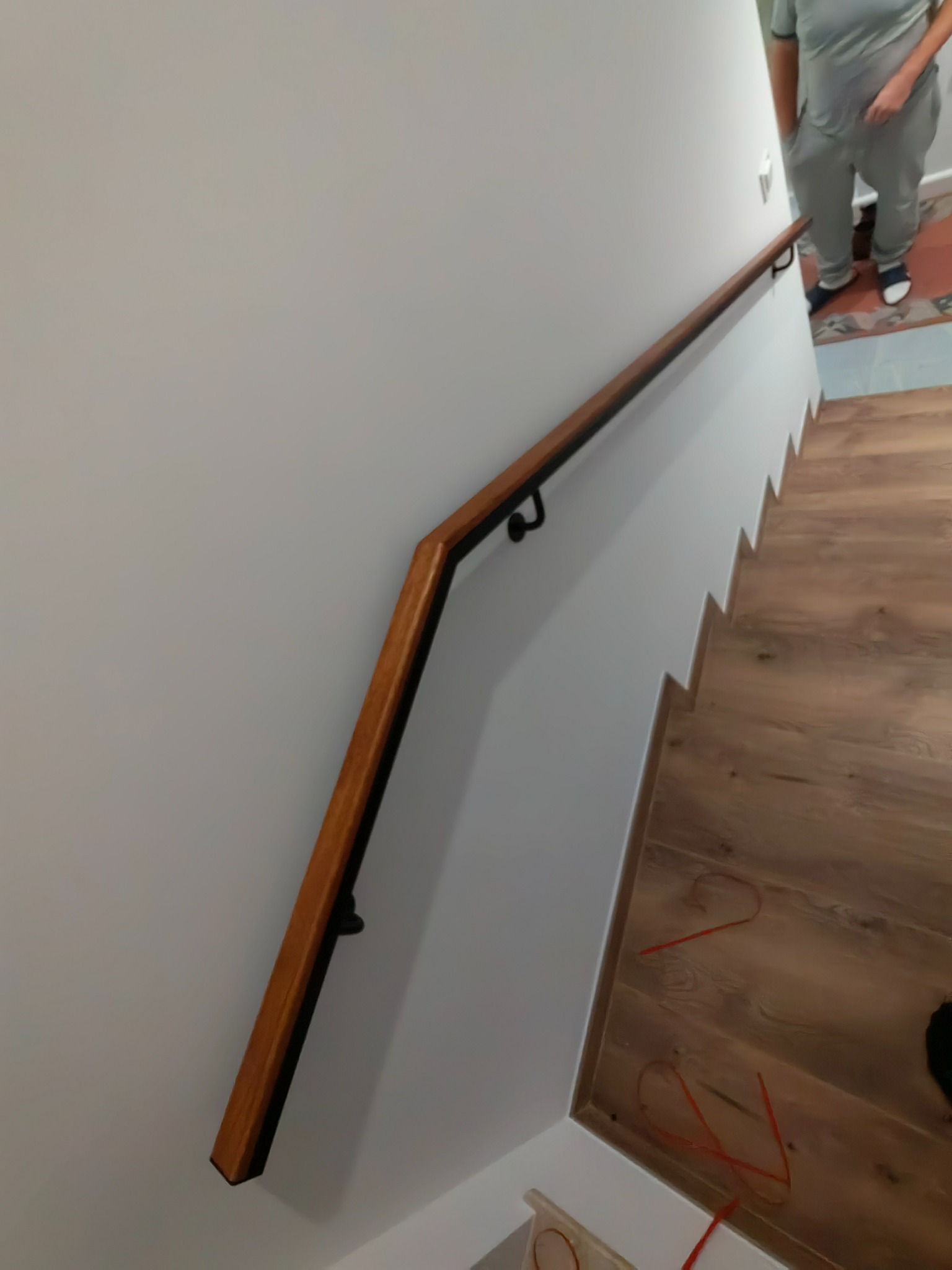 Drewniana poręcz mocowana do ściany wzdłuż schodów, z widocznymi czarnymi uchwytami mocującymi i drewnianą podłogą na schodach. Widoczny fragment osoby na górze schodów.