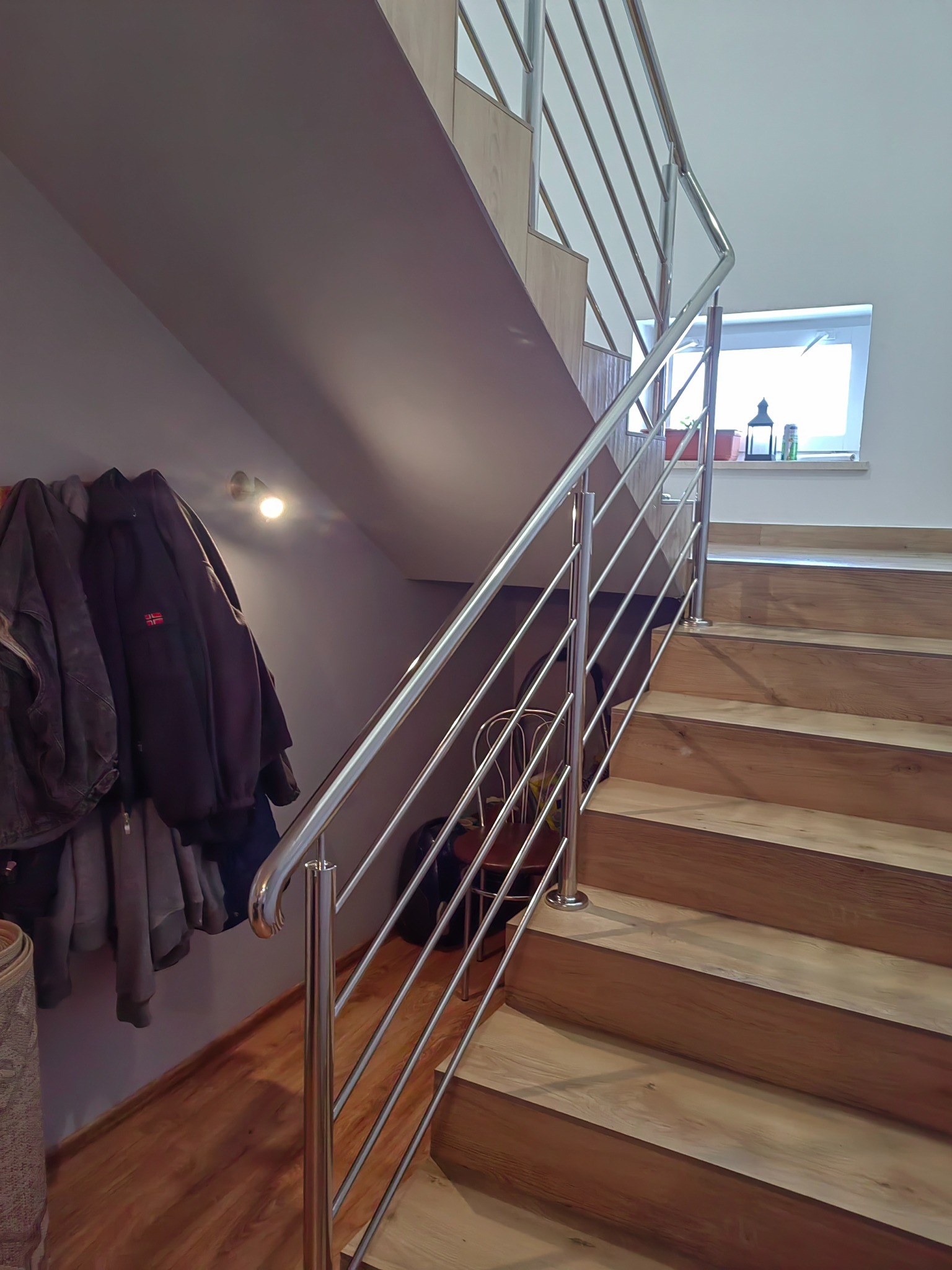 Klatka schodowa z drewnianymi stopniami i nowoczesną, metalową balustradą z wypełnieniem z cienkich prętów. Na ścianie wieszaki z kurtkami. W tle okno z dekoracjami.