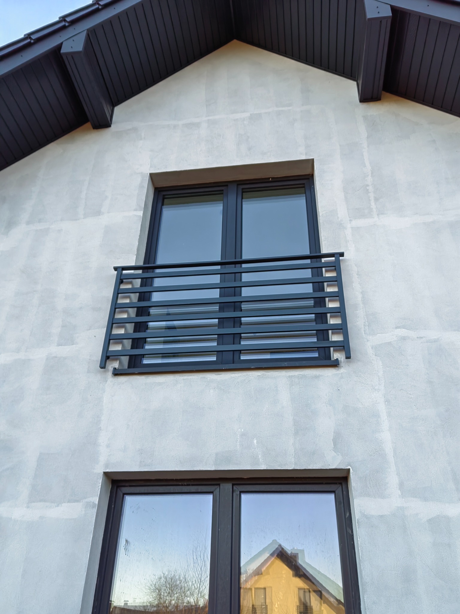 Fasada domu w trakcie wykończenia z oknem balkonowym zabezpieczonym czarną, metalową balustradą o minimalistycznym designie z poziomymi prętami.
