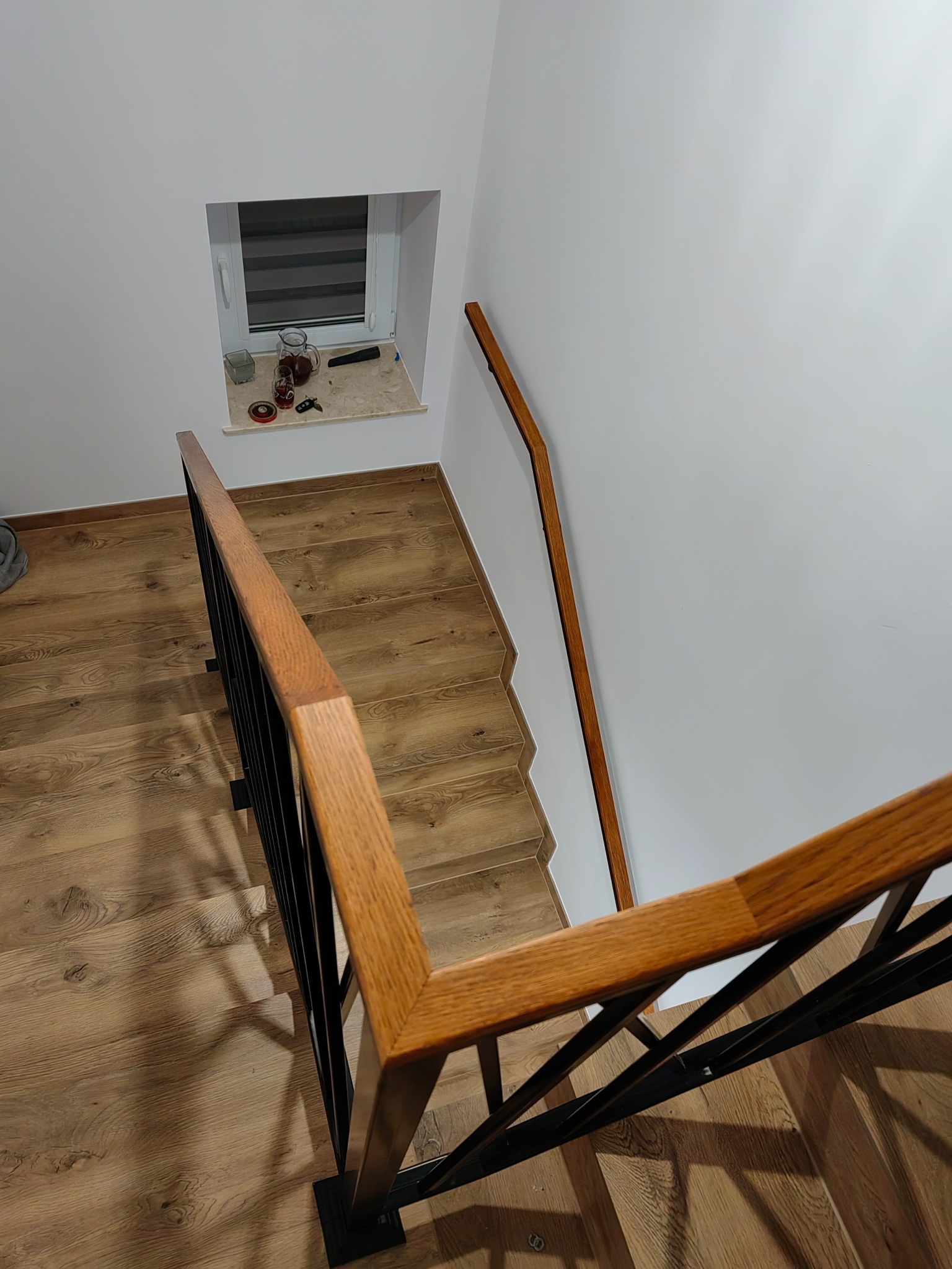 Widok z góry na schody z drewnianymi stopniami i czarną metalową balustradą z drewnianym poręczą; w tle biała ściana z małym oknem z roletami i dekoracjami na parapecie.