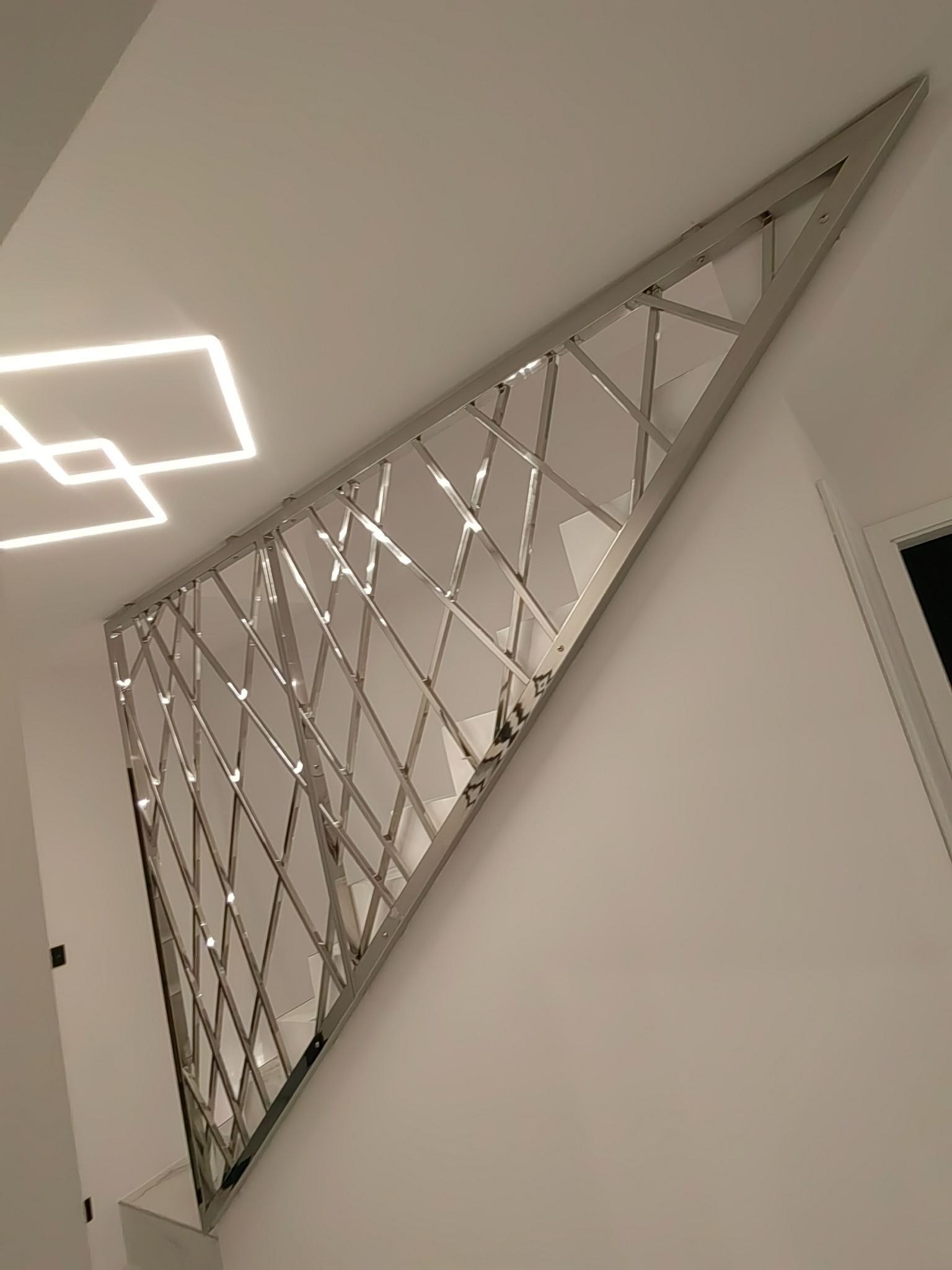 Nietypowa balustrada schodowa ze stali nierdzewnej w kształcie trójkąta z ukośnymi poprzeczkami, nowoczesne oświetlenie sufitowe w tle.