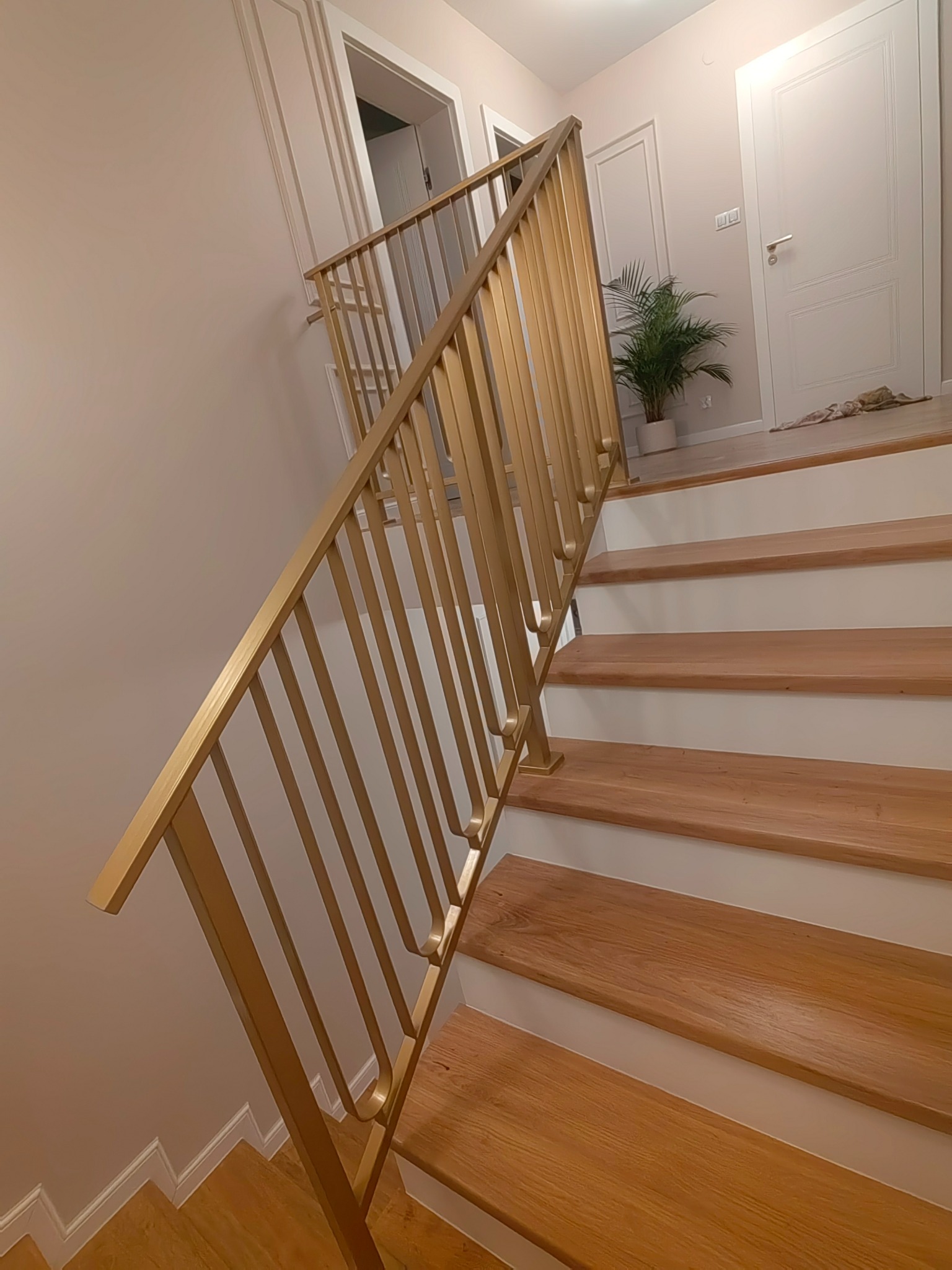 Złota, metalowa poręcz schodowa z pionowymi szczeblami, zamontowana na drewnianych schodach z białymi podstopnicami, w tle widoczne drzwi i roślina doniczkowa.