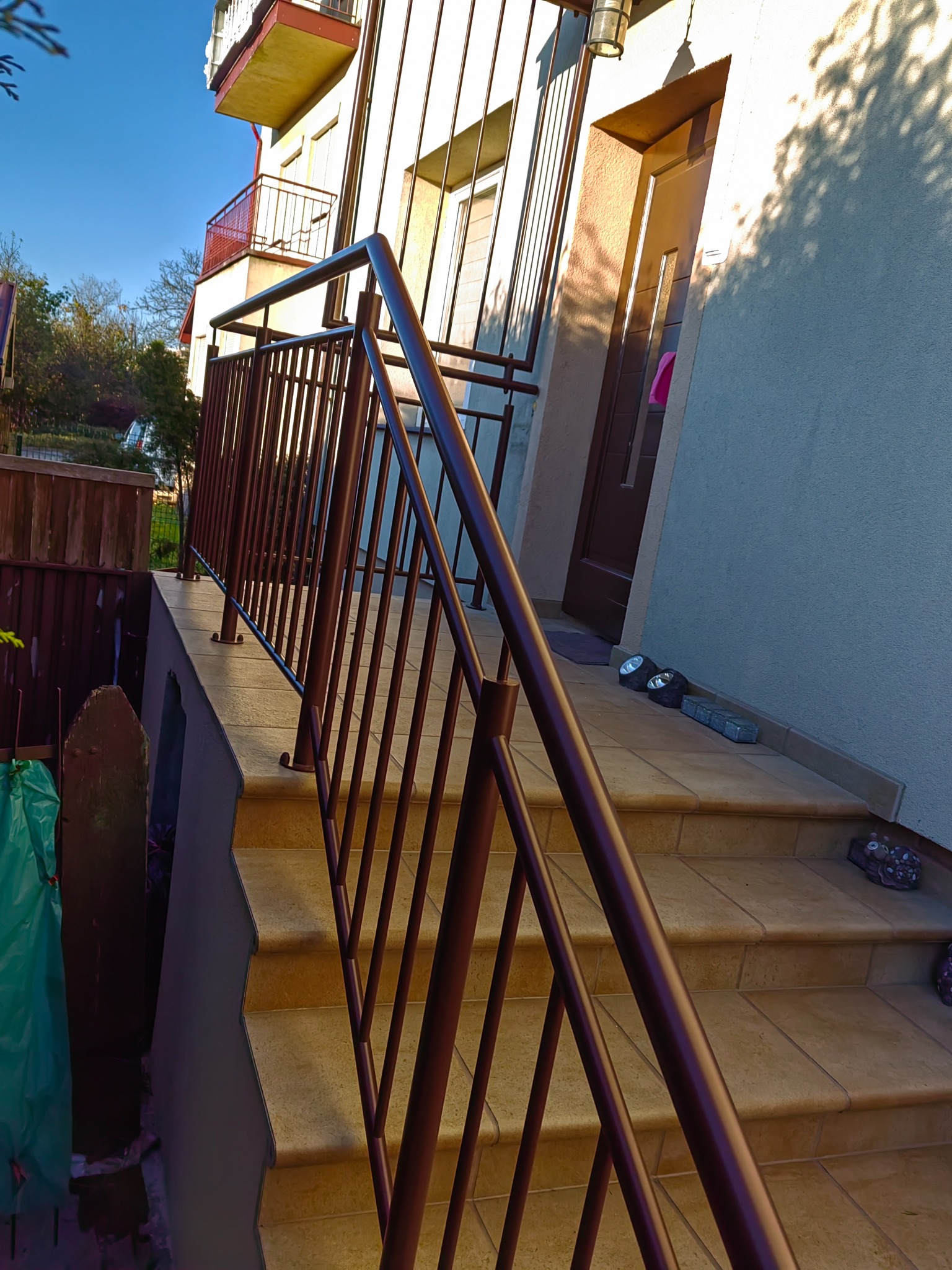 Brązowa metalowa balustrada przy schodach wejściowych do budynku jednorodzinnego, widok z dołu na balustradę i część elewacji.