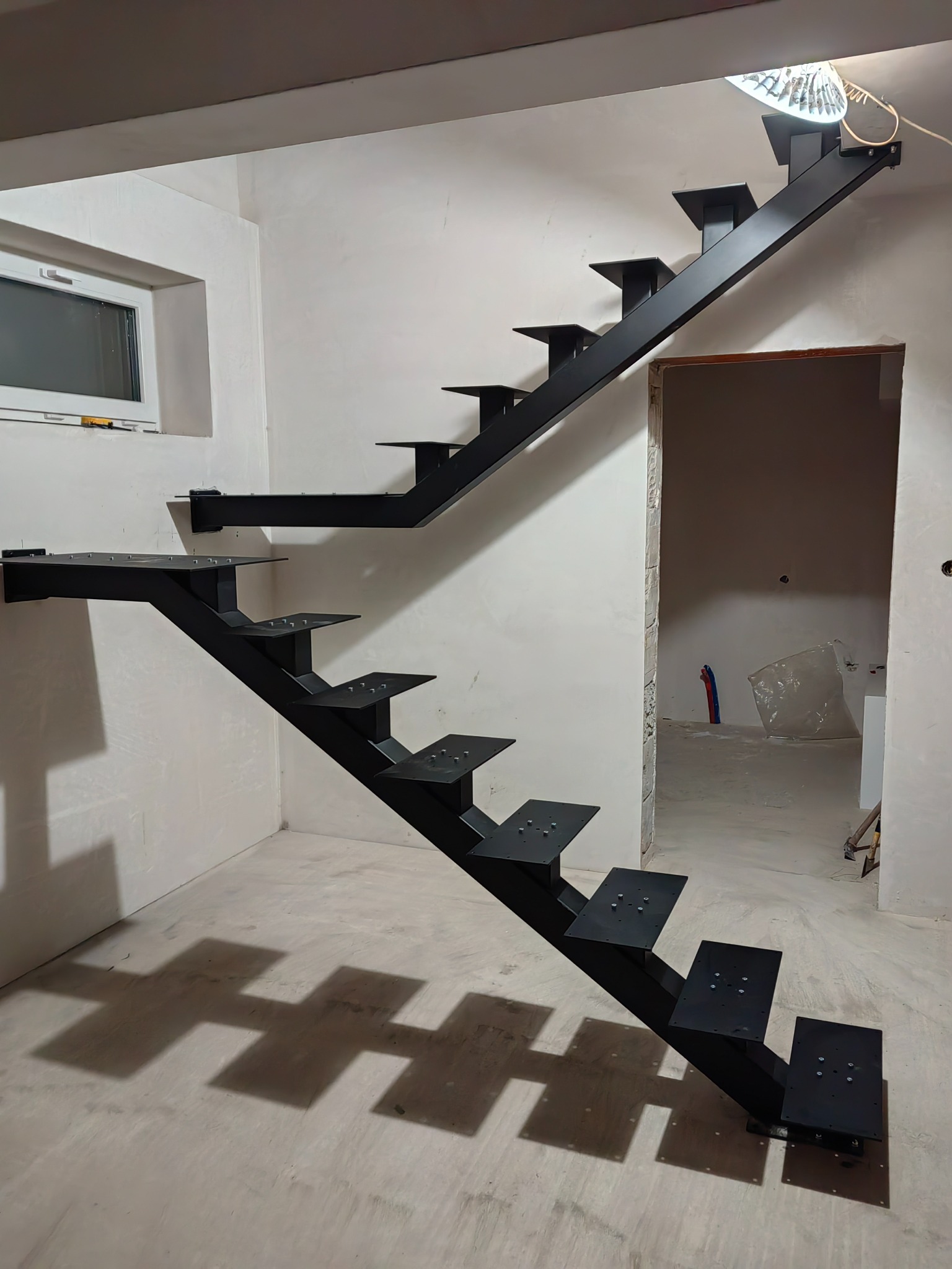Surowe, czarne schody metalowe bez poręczy, zamontowane w surowym wnętrzu z widocznym oknem i przejściem do innego pomieszczenia w trakcie wykończenia.