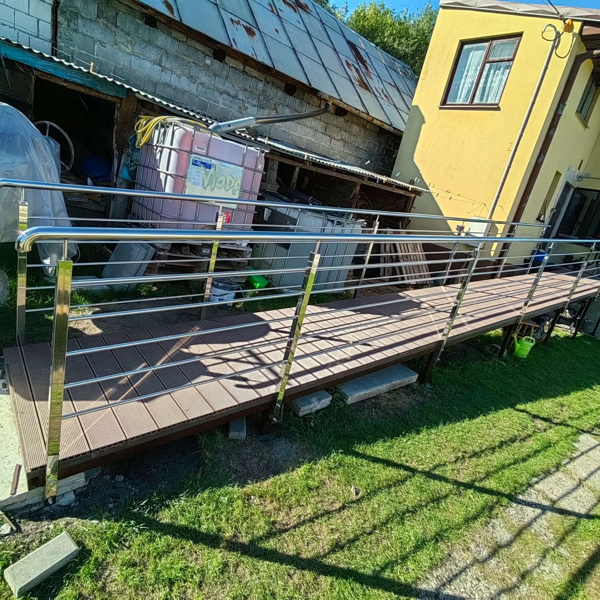 Zewnętrzny podest z drewnopodobnych desek z chromowanymi barierkami, widok na budynek gospodarczy i żółty dom jednorodzinny.