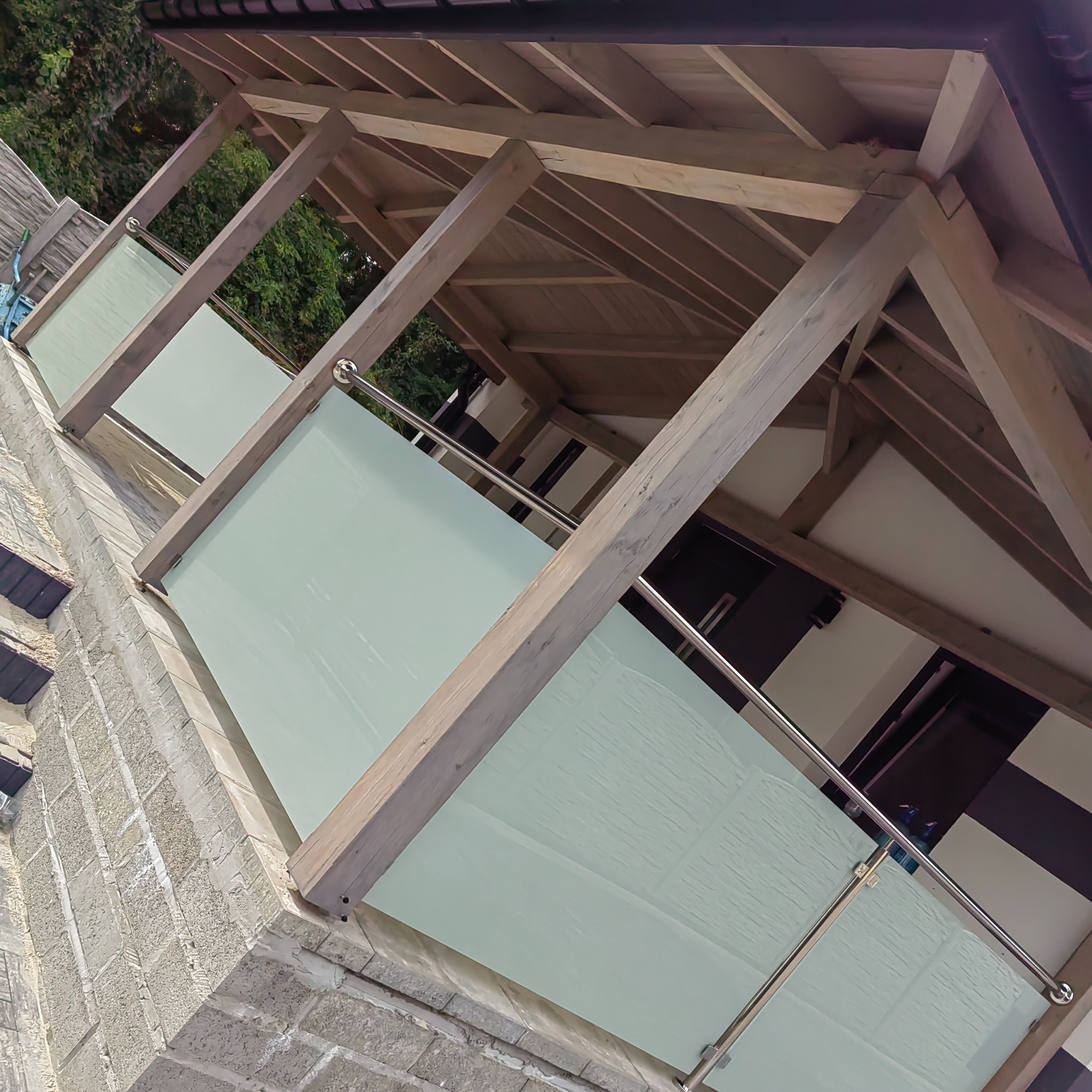 Zadaszony balkon z drewnianą konstrukcją i matowymi szklanymi panelami, widok z dołu na elewację budynku.