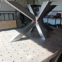 Surowa, metalowa podstawa stołu z centralnym węzłem spawów, umieszczona na perforowanym stole warsztatowym w jasnym warsztacie.