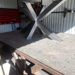 Surowa, spawana podstawa stołu w kształcie gwiazdy z profili stalowych, na stole spawalniczym w warsztacie, widoczne narzędzia i pojemniki z akcesoriami na ścianie.