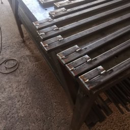 Spawalnia - Metalowe pręty z przyspawanymi elementami leżące na stole warsztatowym, prawdopodobnie elementy konstrukcji ogrodzenia lub bramy w trakcie produkcji.
