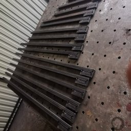 Spawalnia - Metalowe pręty z przyspawanymi elementami leżące na metalowym, perforowanym stole warsztatowym, przygotowane do dalszej obróbki lub montażu.