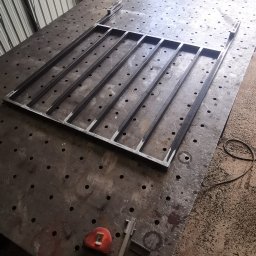 Spawalnia - Metalowa konstrukcja w trakcie spawania na stole warsztatowym, widoczny spawany fragment bramy lub ogrodzenia, miarka i suwmiarka obok.