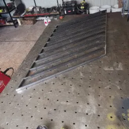 Konstrukcja metalowa bramy wjazdowej w trakcie spawania na stole spawalniczym, widoczne narzędzia ślusarskie i pojemnik ze stopiwem spawalniczym.