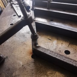 Spawalnia - Metalowy profil zespawany pod kątem prostym, przytrzymywany ściskami stolarskimi na metalowym stole warsztatowym z widocznymi otworami.