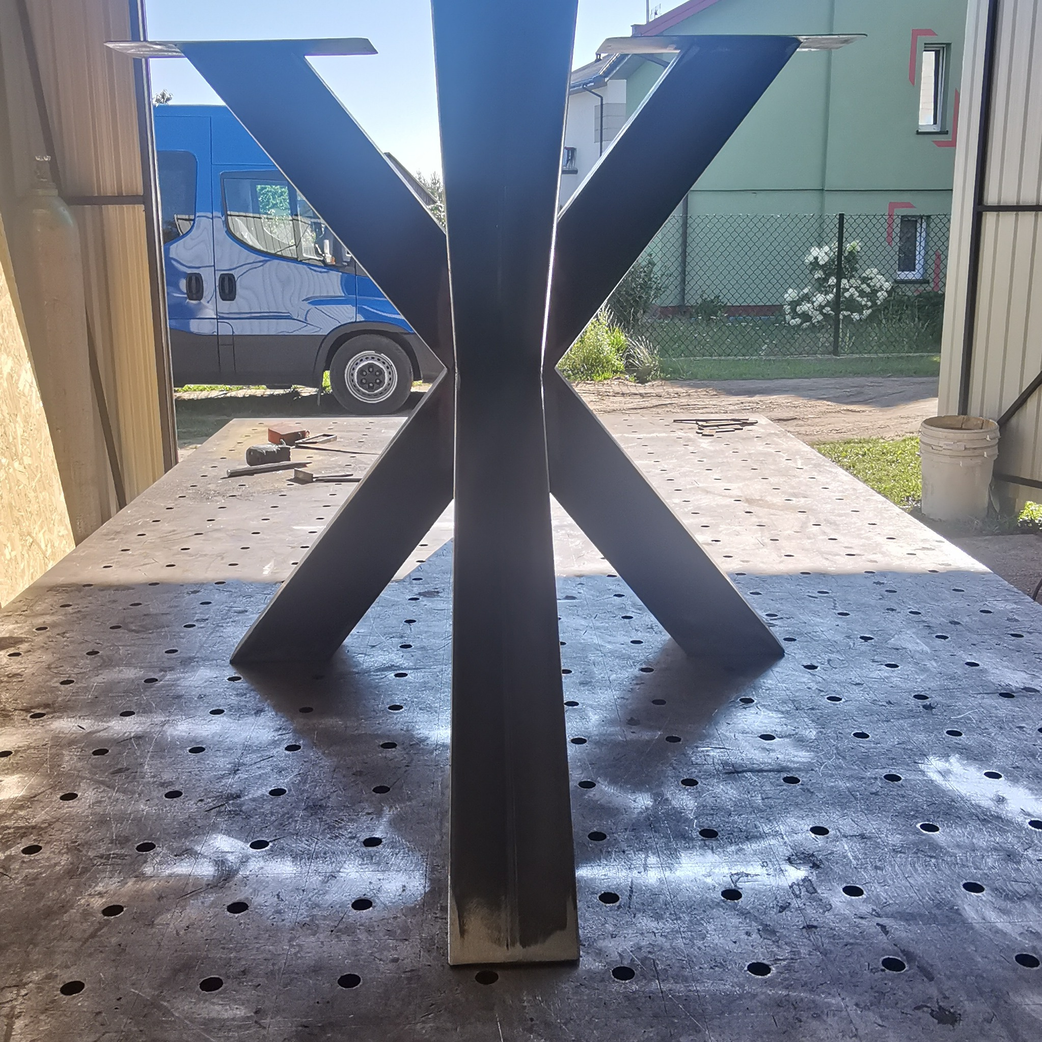 Metalowa podstawa stołu w kształcie litery X, spawana na stole spawalniczym, w tle budynek i samochód dostawczy.
