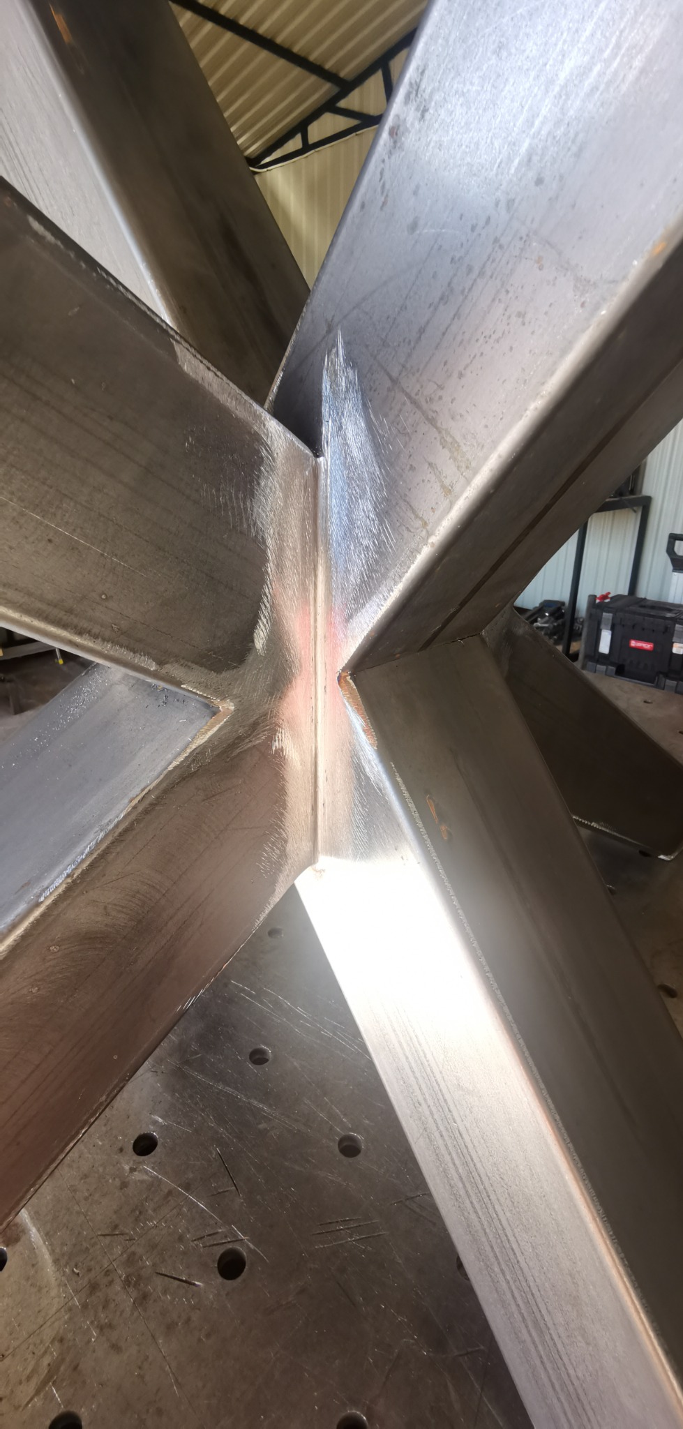 Surowe, metalowe elementy konstrukcyjne zespawane w kształt gwiazdy, leżące na perforowanym stole spawalniczym, widoczne ślady obróbki i spawania.