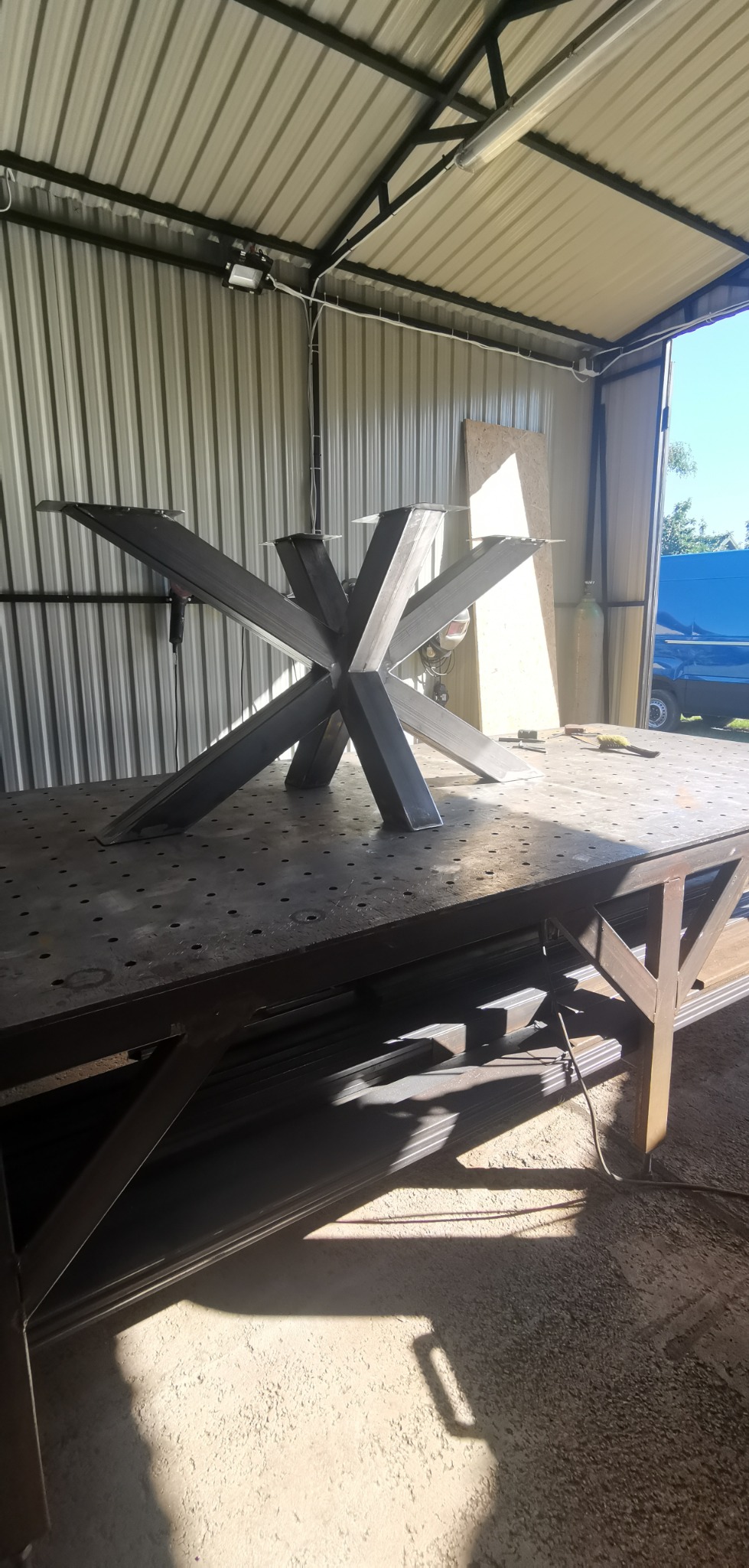 Metalowa podstawa stołu w kształcie gwiazdy, spawana na stole warsztatowym w hali z blachy falistej, z widocznymi narzędziami i materiałami.