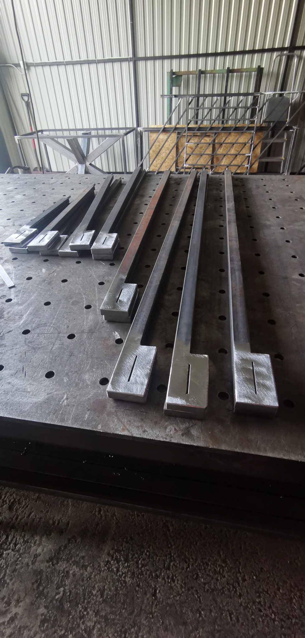 Surowe, metalowe elementy z wyciętymi otworami ułożone na perforowanym stole warsztatowym w hali produkcyjnej.