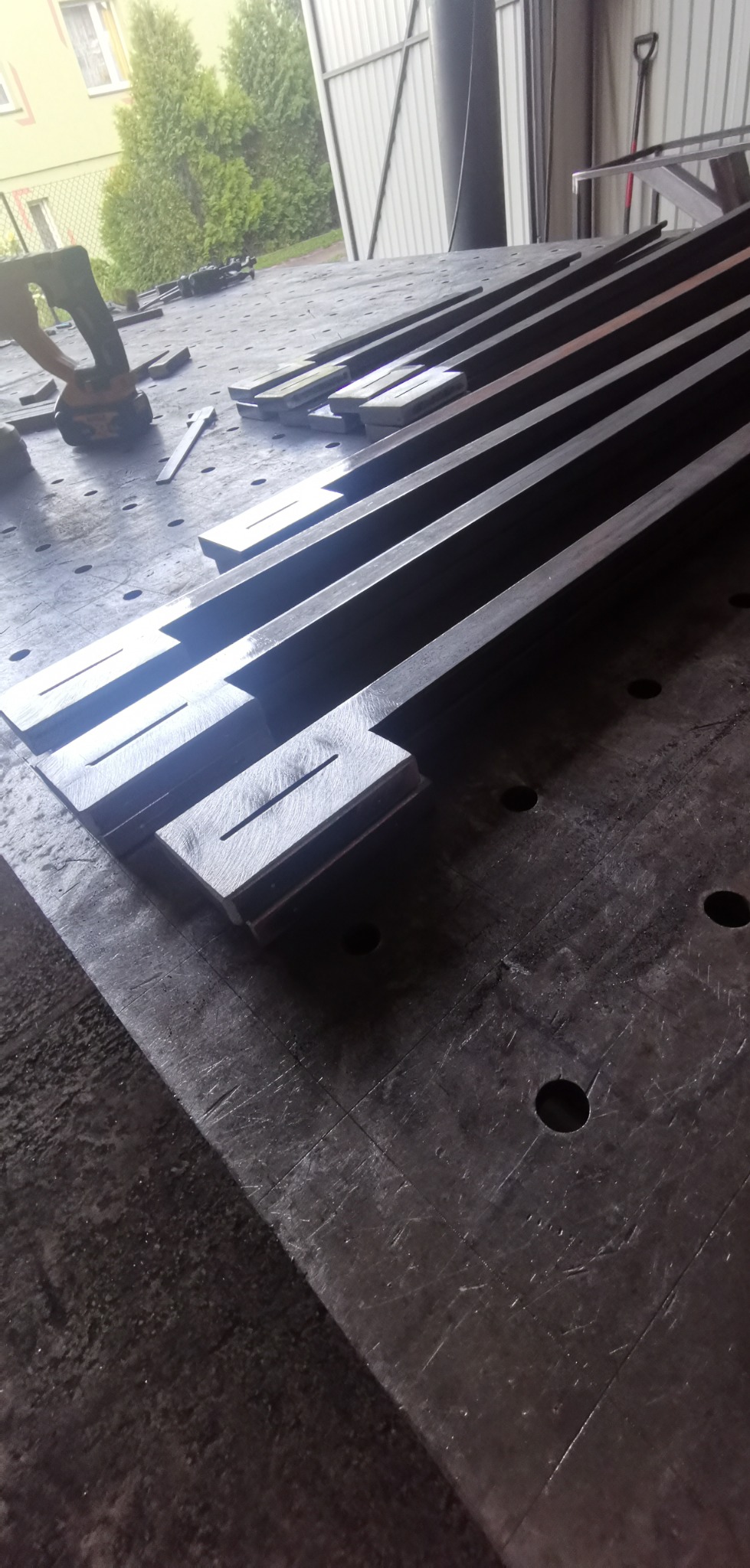 Przygotowane do spawania metalowe profile z wyciętymi otworami, ułożone na perforowanym stole spawalniczym, z widocznym narzędziem akumulatorowym w tle.