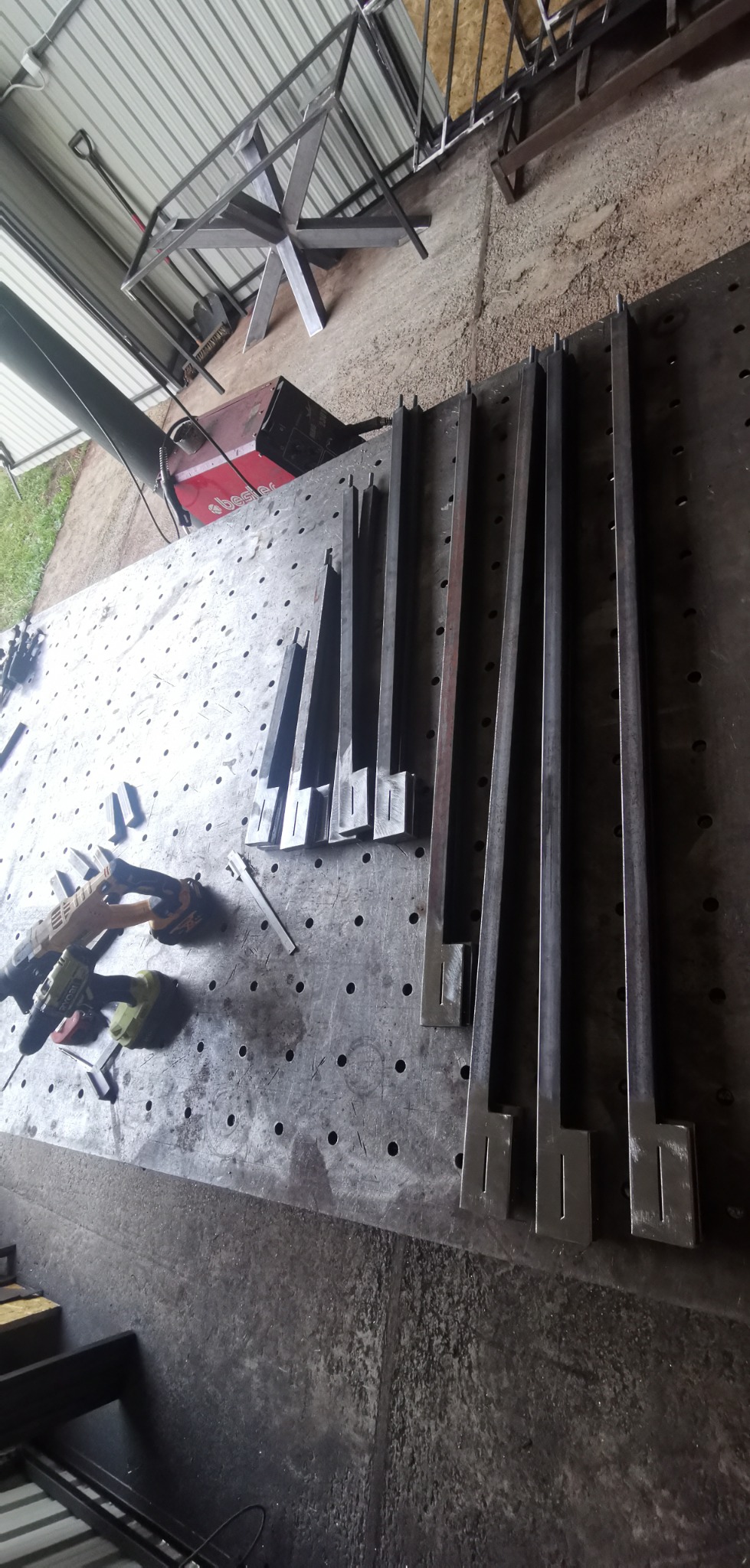 Przygotowane elementy metalowe z wyciętymi otworami, leżące na perforowanym stole warsztatowym obok wiertarek akumulatorowych i spawarki, gotowe do dalszej obróbki i montażu konstrukcji.