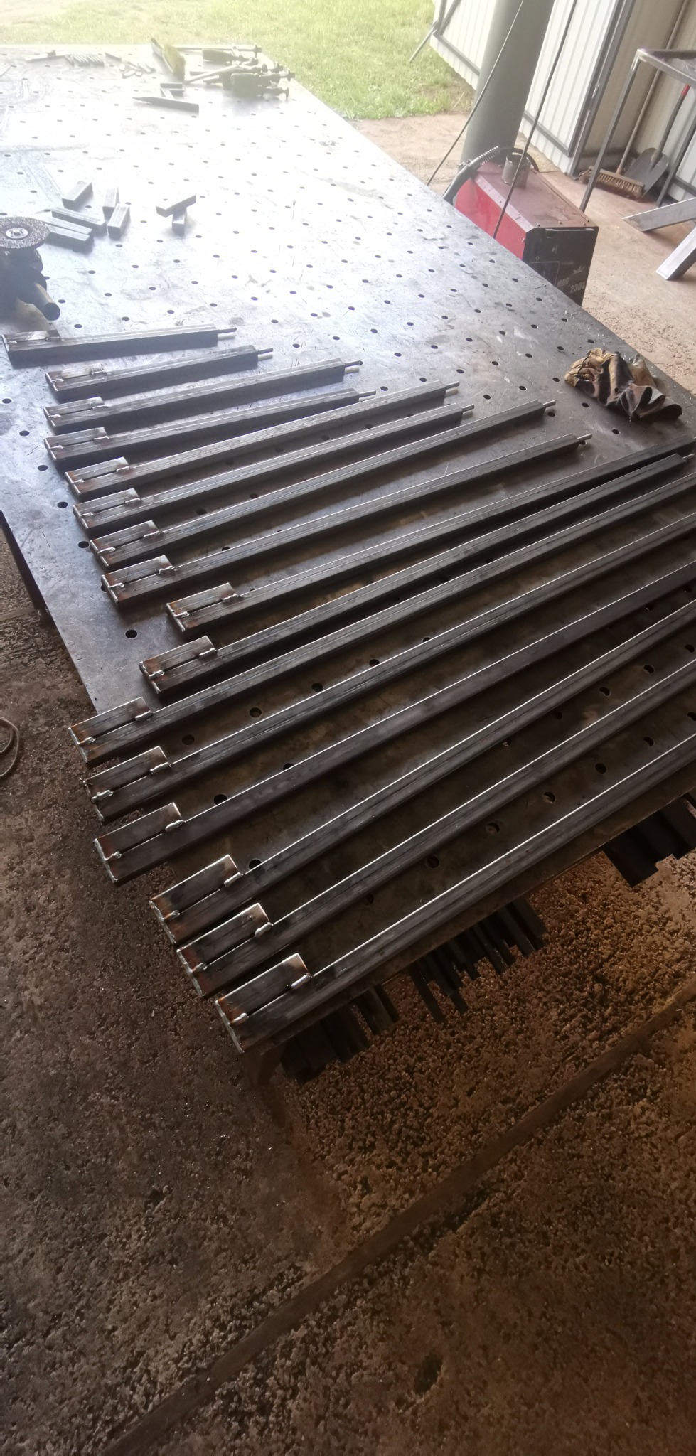 Metalowe profile z przyspawanymi elementami ułożone równolegle na perforowanym stole warsztatowym w hali, widoczne narzędzia i rękawice robocze.