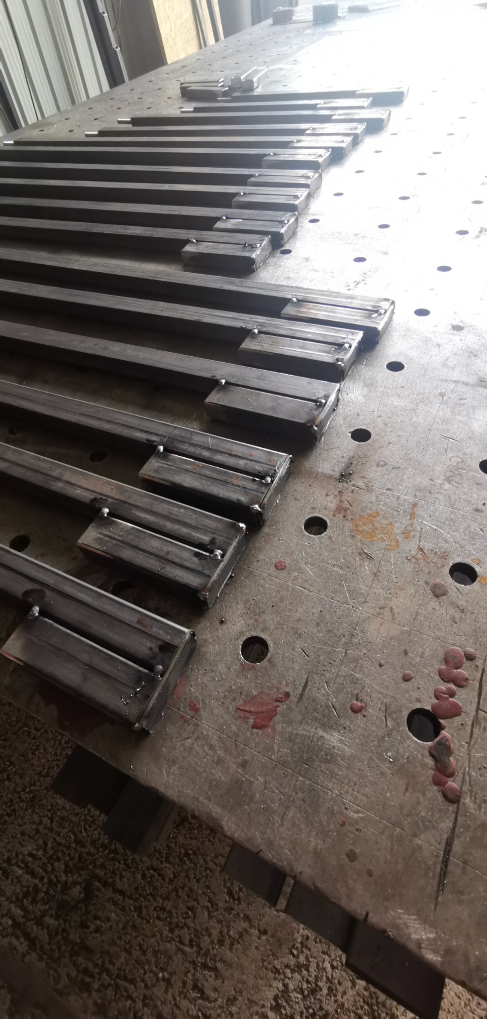 Metalowe profile kwadratowe, spawane z krótkimi elementami, ułożone rzędami na metalowym stole spawalniczym z widocznymi śladami użytkowania i otworami.