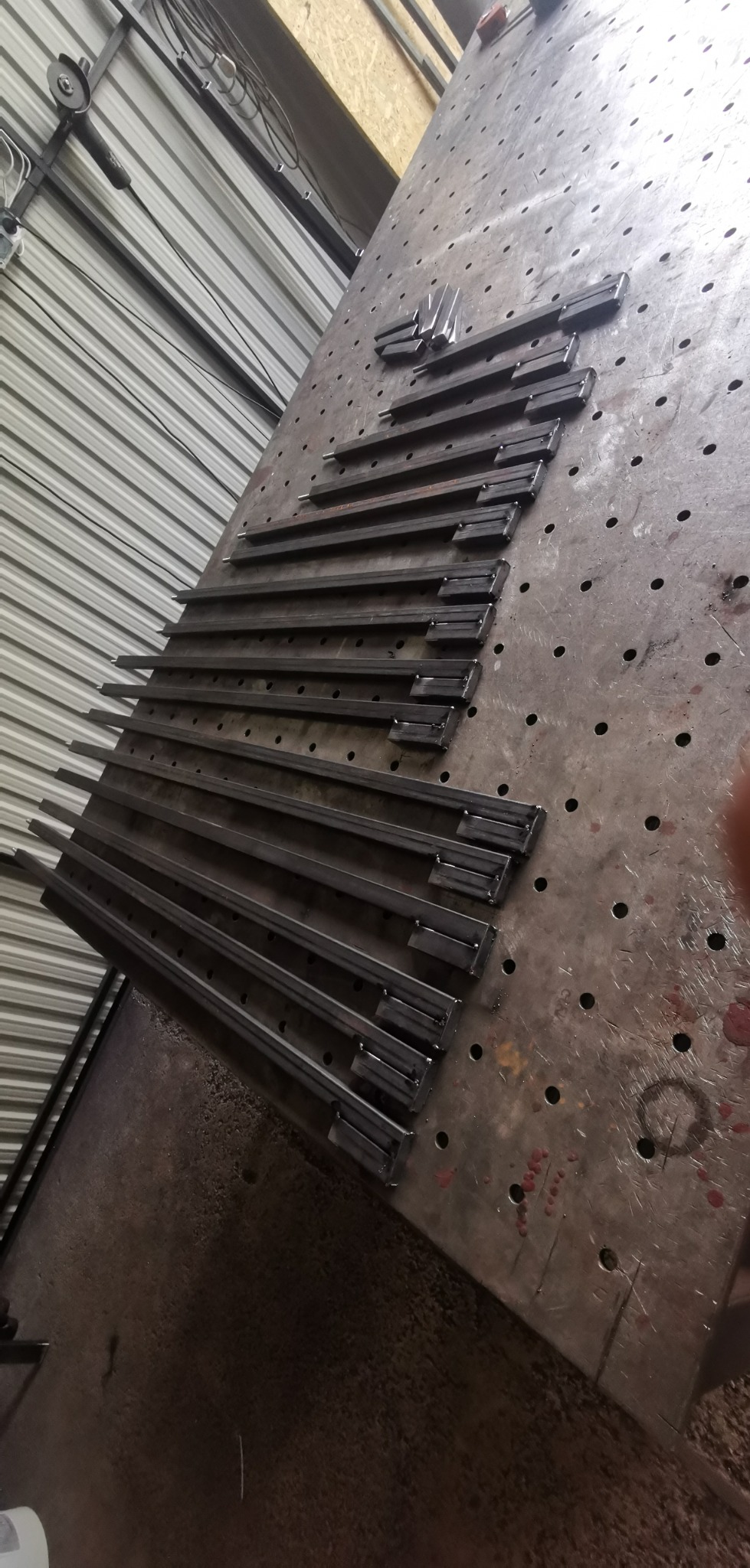 Metalowe pręty z przyspawanymi elementami leżące na metalowym, perforowanym stole warsztatowym, przygotowane do dalszej obróbki lub montażu.