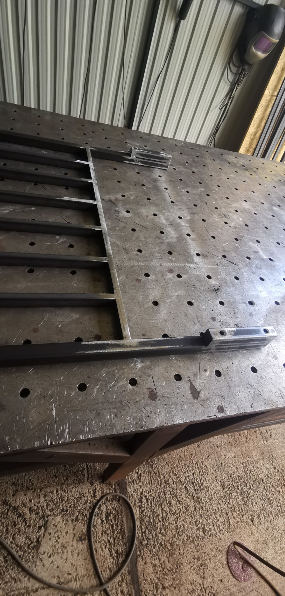 Metalowa konstrukcja przypominająca fragment ogrodzenia z prętów, w trakcie spawania na stole spawalniczym z widocznymi otworami, w tle maska spawalnicza.