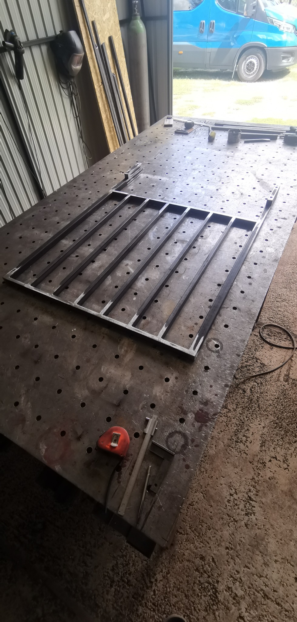 Metalowa konstrukcja w trakcie spawania na stole warsztatowym, widoczny spawany fragment bramy lub ogrodzenia, miarka i suwmiarka obok.