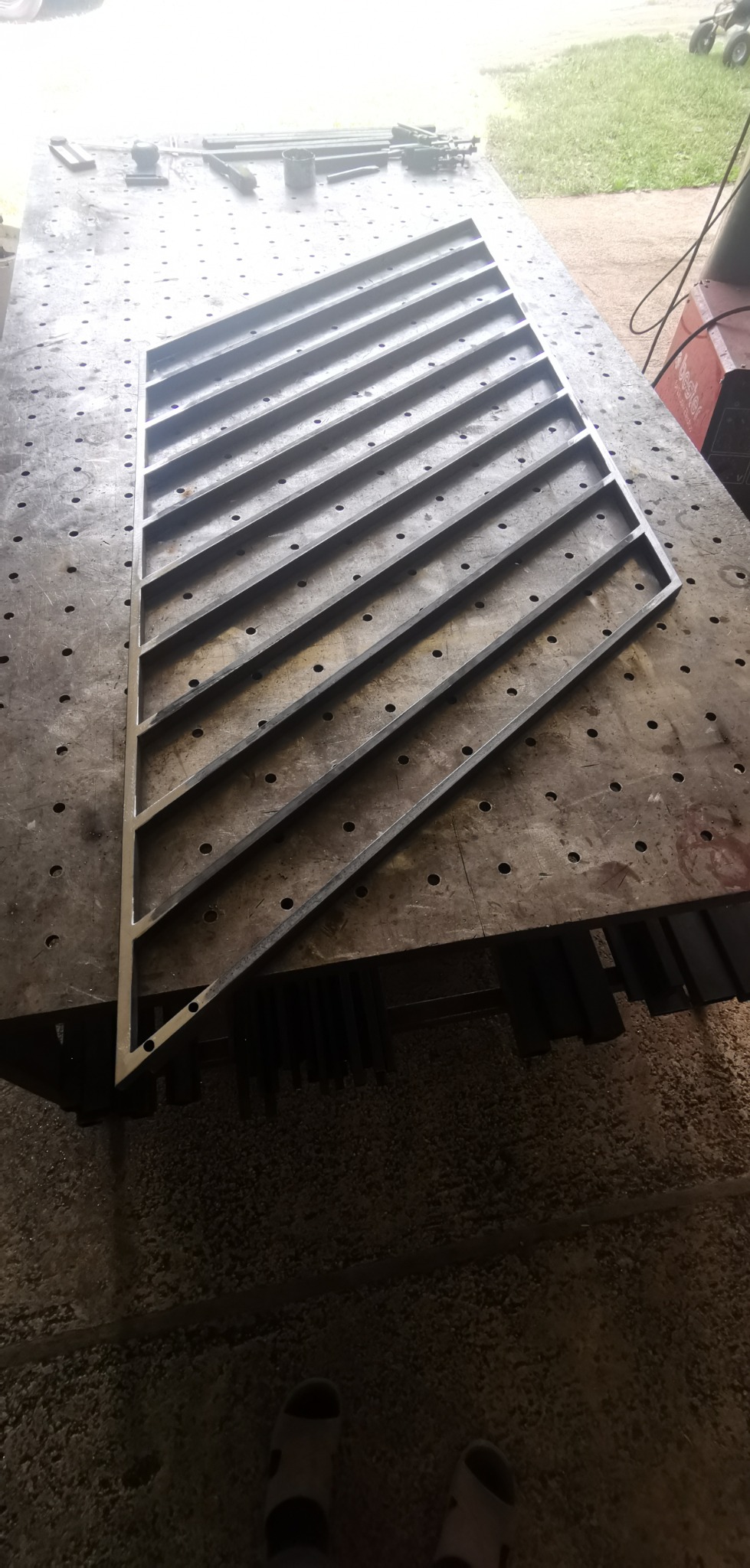 Metalowa konstrukcja z ukośnymi prętami, prawdopodobnie fragment bramy lub ogrodzenia, leży na stole spawalniczym z widocznymi otworami i narzędziami w tle.