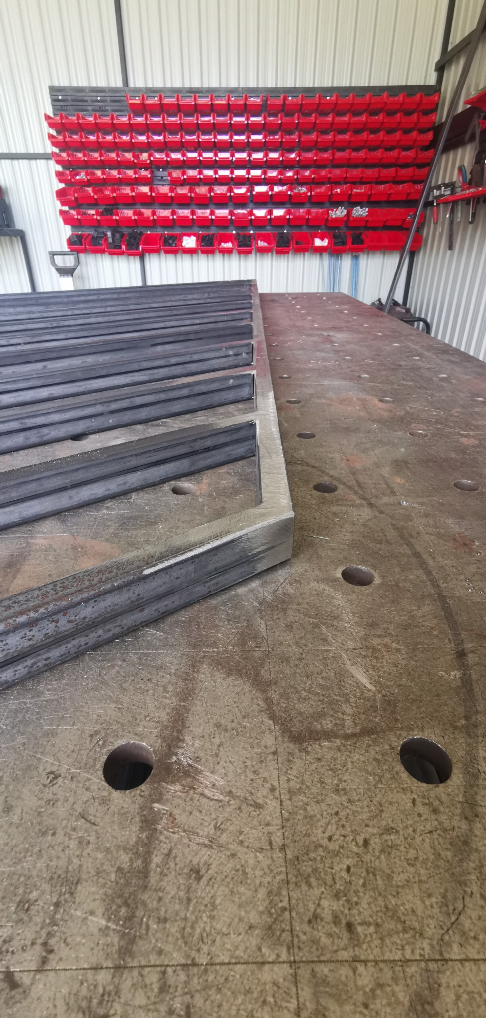 Metalowe profile ułożone na stole spawalniczym z perforacją, w tle widoczne czerwone pojemniki na narzędzia.