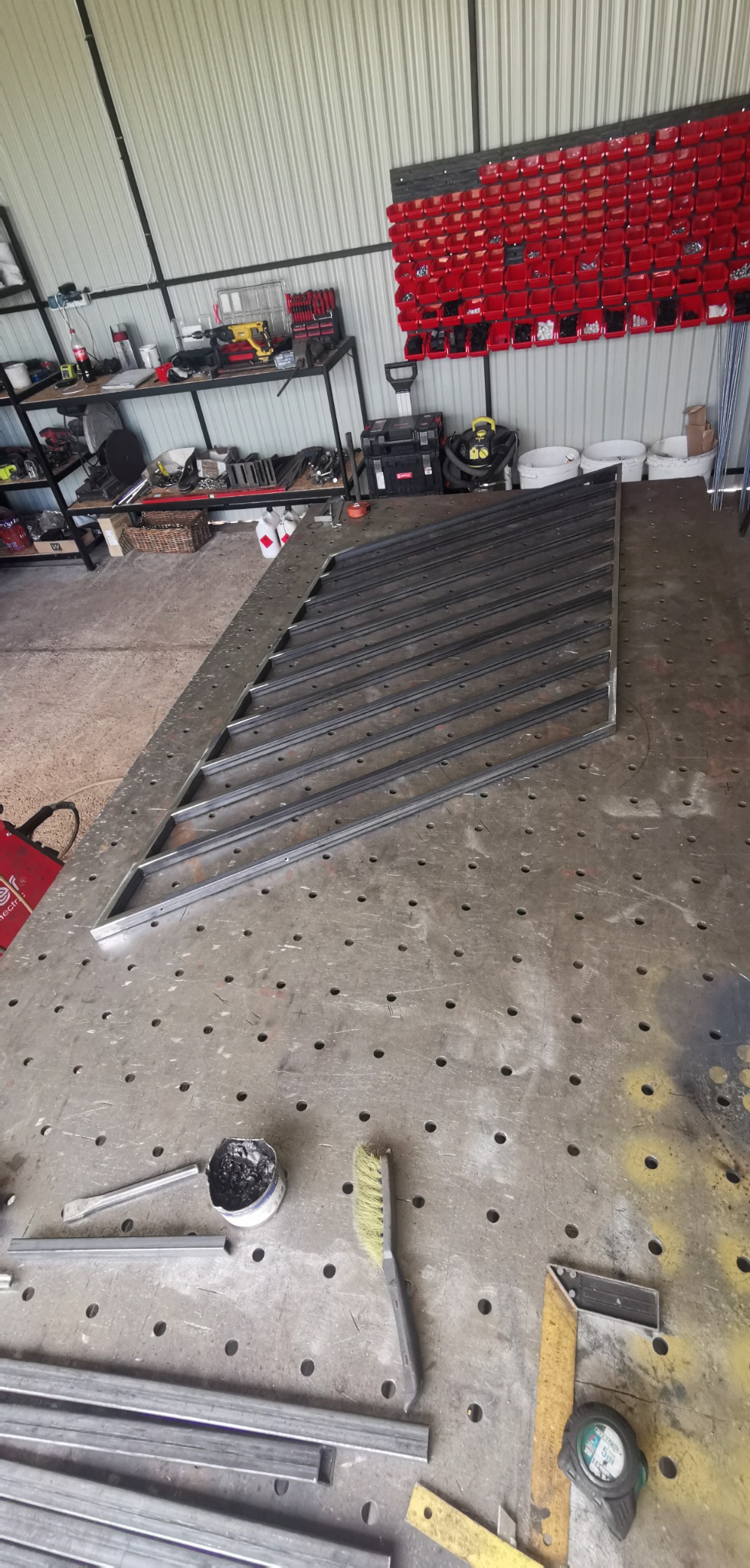 Konstrukcja metalowa bramy wjazdowej w trakcie spawania na stole spawalniczym, widoczne narzędzia ślusarskie i pojemnik ze stopiwem spawalniczym.