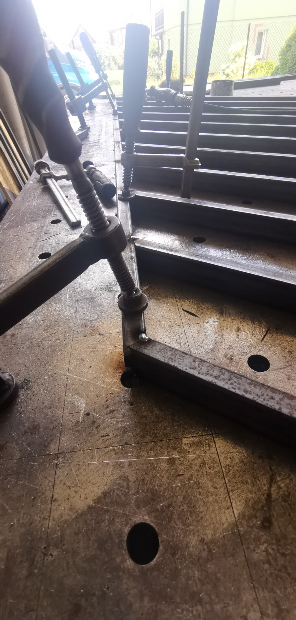 Metalowy profil zespawany pod kątem prostym, przytrzymywany ściskami stolarskimi na metalowym stole warsztatowym z widocznymi otworami.