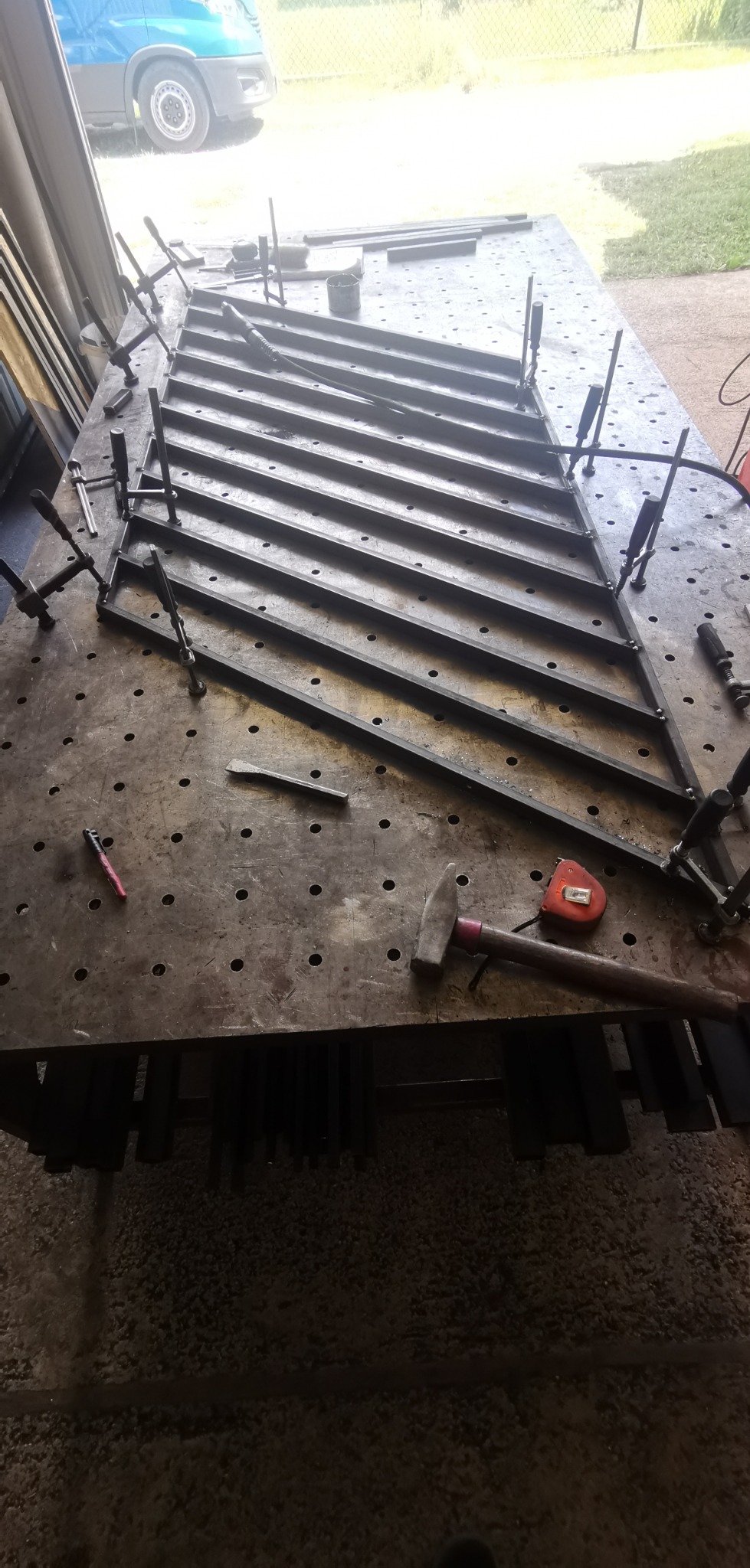 Metalowa konstrukcja kratowa w trakcie spawania, mocowana zaciskami na perforowanym stole warsztatowym, obok leżą młotek i miarka.