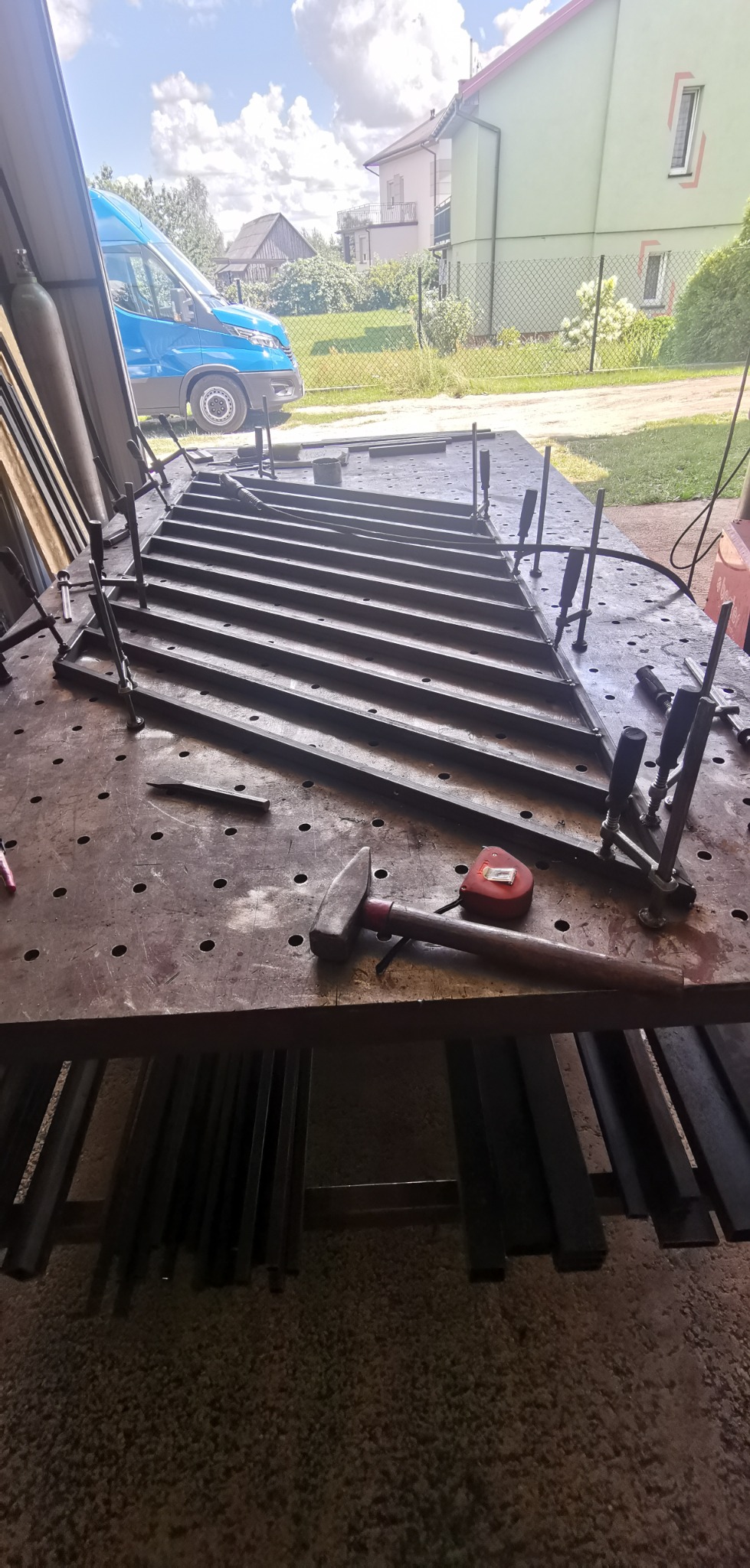 Konstrukcja metalowa z poprzeczkami, prawdopodobnie element bramy, mocowana klamrami na perforowanym stole warsztatowym, obok leżą narzędzia: młotek i miara zwijana, w tle widoczny samochód...