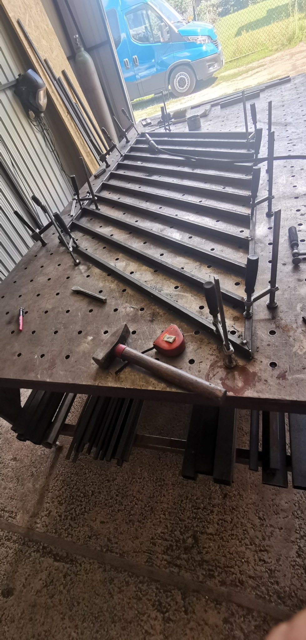 Spawanie konstrukcji metalowej w warsztacie w Skierniewicach. Widoczny stół spawalniczy z przyrządami, młotek, miarka, klamry śrubowe mocujące elementy, oraz fragment niebieskiego samochodu...