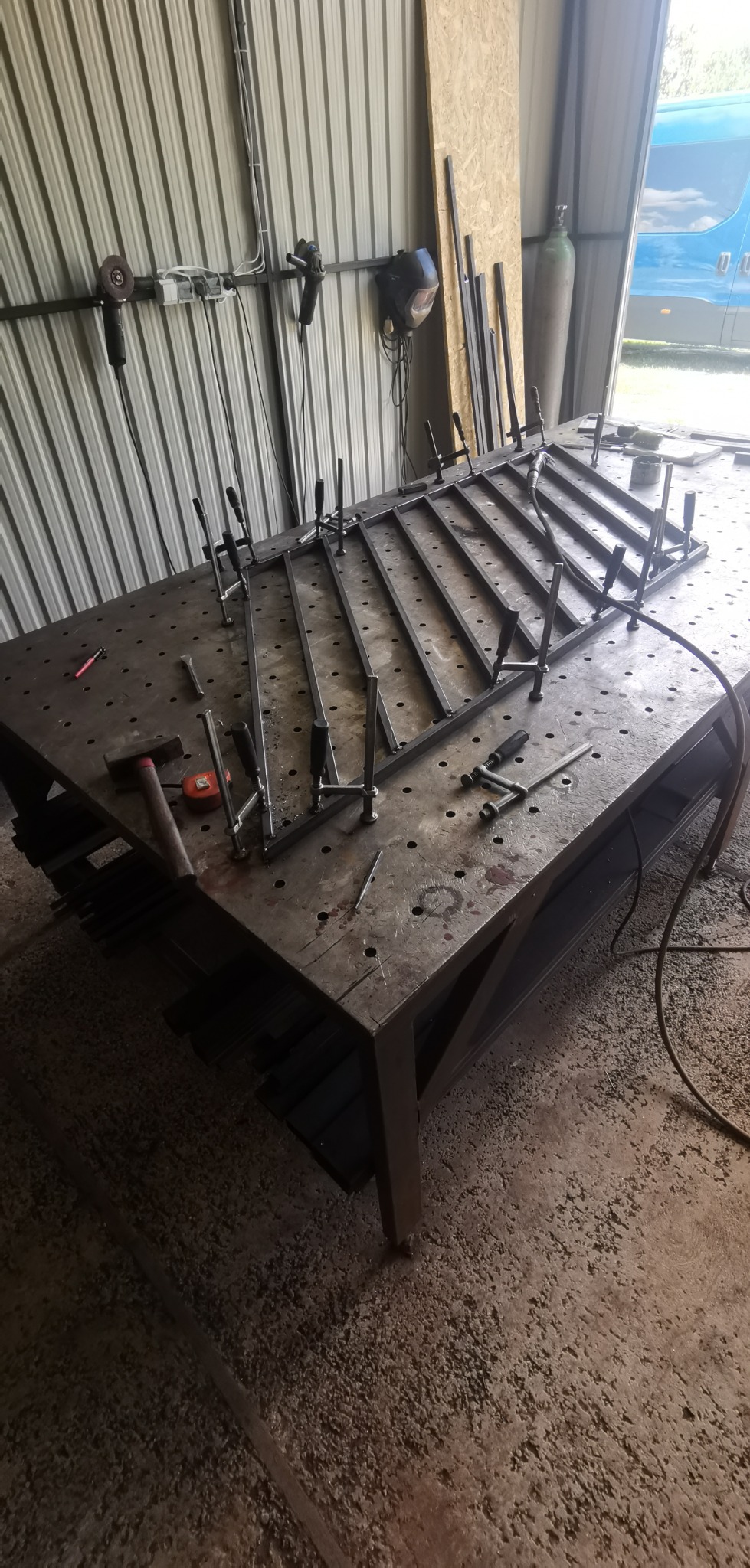 Metalowa konstrukcja schodkowa w trakcie spawania na stole spawalniczym, zabezpieczona ściskami, w tle ściana z blachy falistej i narzędzia.