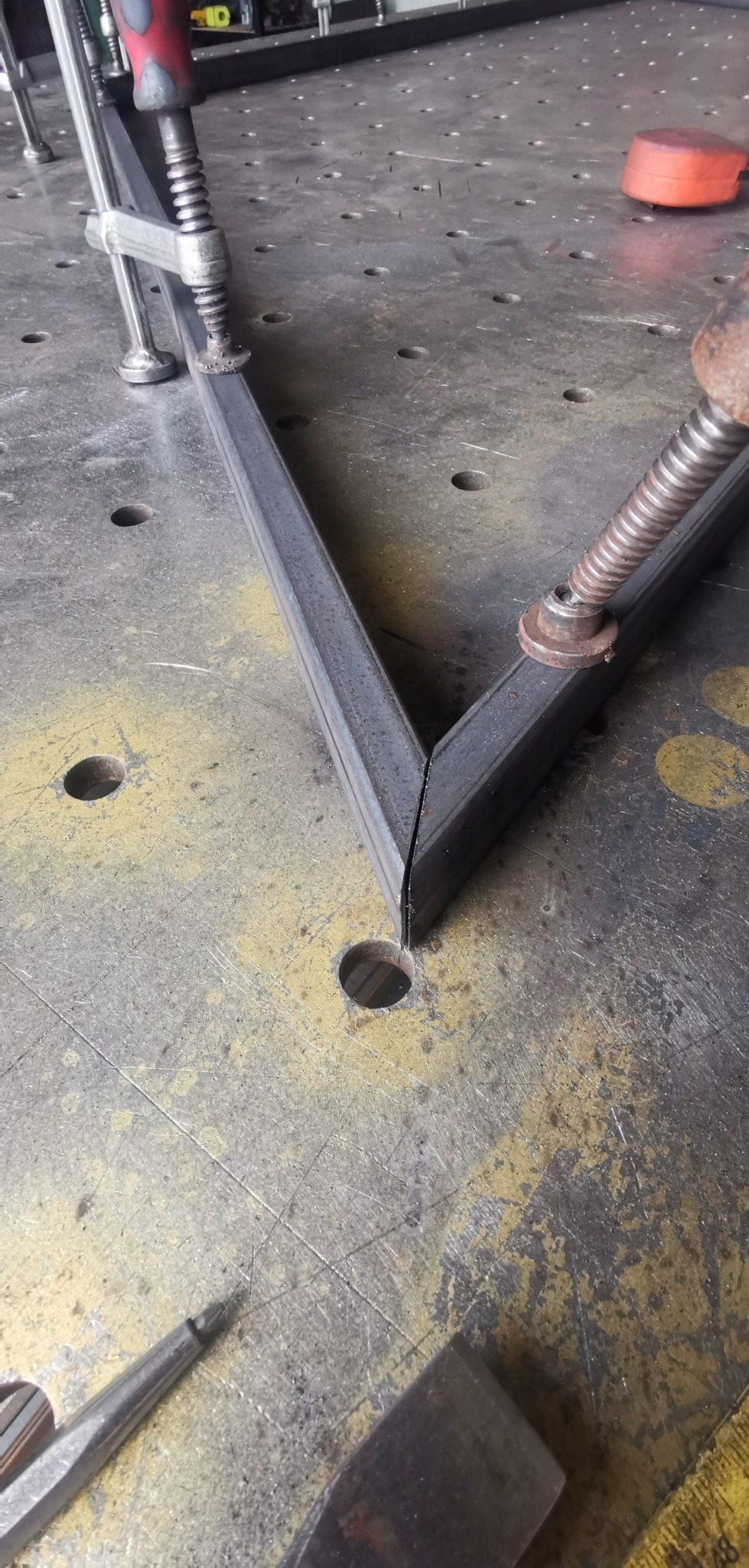 Dwa metalowe profile kątowe, zaciśnięte razem na perforowanym stole spawalniczym za pomocą ścisków stolarskich, przygotowane do spawania narożnika pod kątem prostym. Widoczne narzędzia i ślady po...