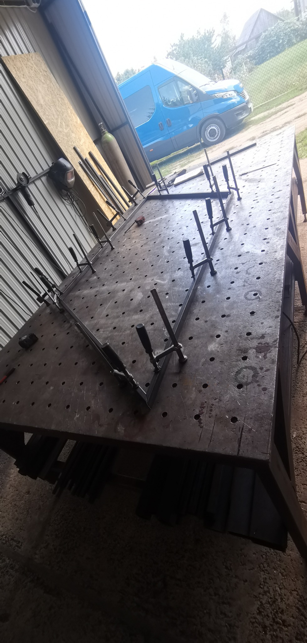 Metalowe profile mocowane klamrami na perforowanym stole spawalniczym w warsztacie, w tle butla z gazem i niebieski samochód dostawczy.