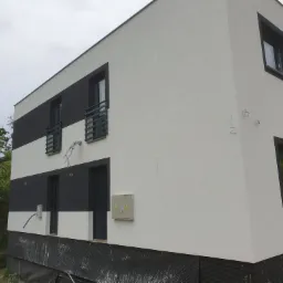 Nowoczesny budynek z biało-czarną elewacją, widoczne okna z barierkami, skrzynka elektryczna na ścianie, oraz wystające kable i przewody w pobliżu górnych okien.