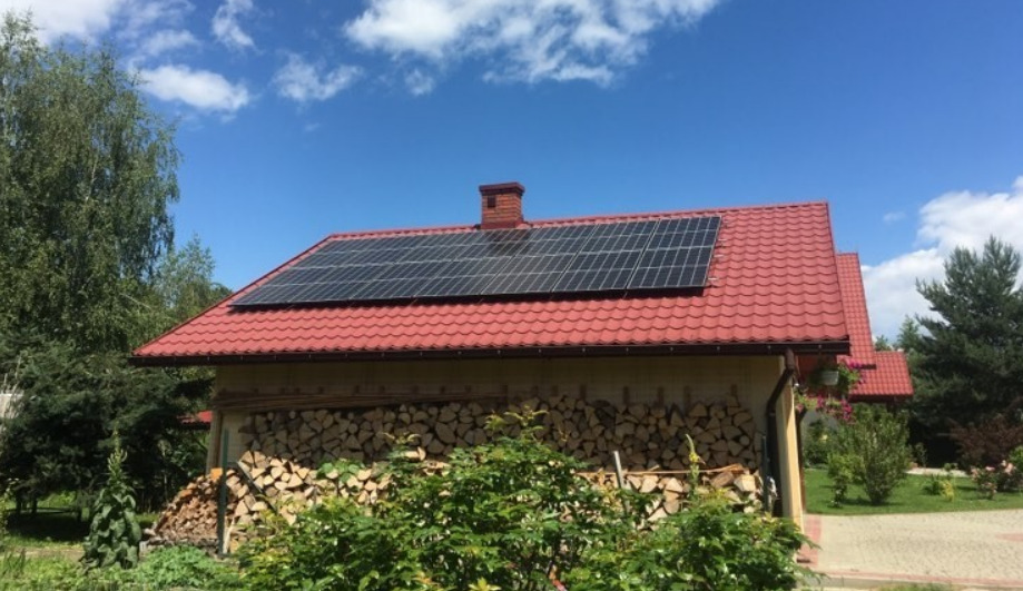 Czerwony dach z panelami słonecznymi na parterowym domu z ułożonym drewnem na ścianie, błękitne niebo w tle.